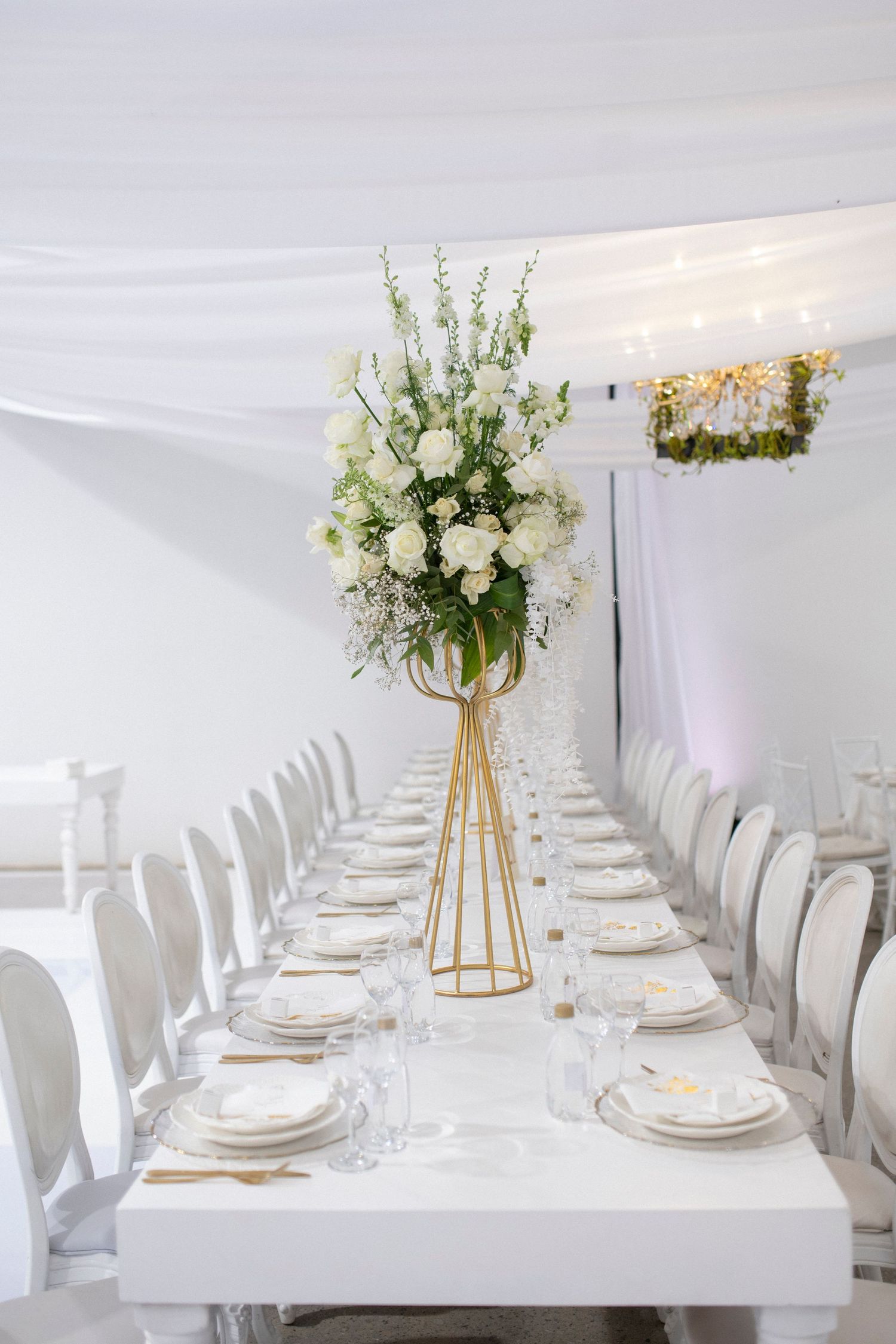 White wedding table setting with tall gold geometric vase holding white and green floral arrangement.