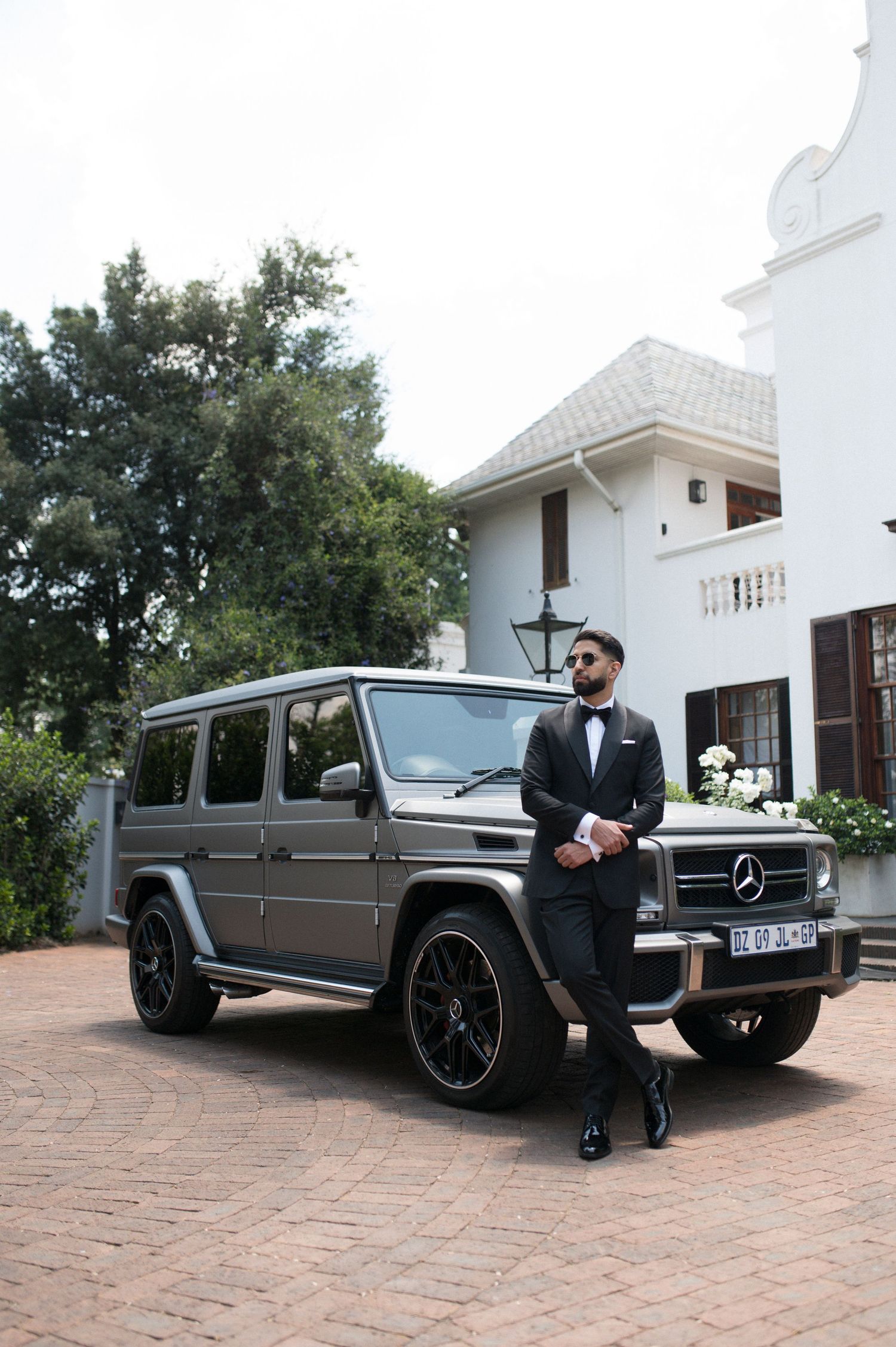 A luxurious grey Mercedes G-Wagon parked in front of a white colonial-style mansion.