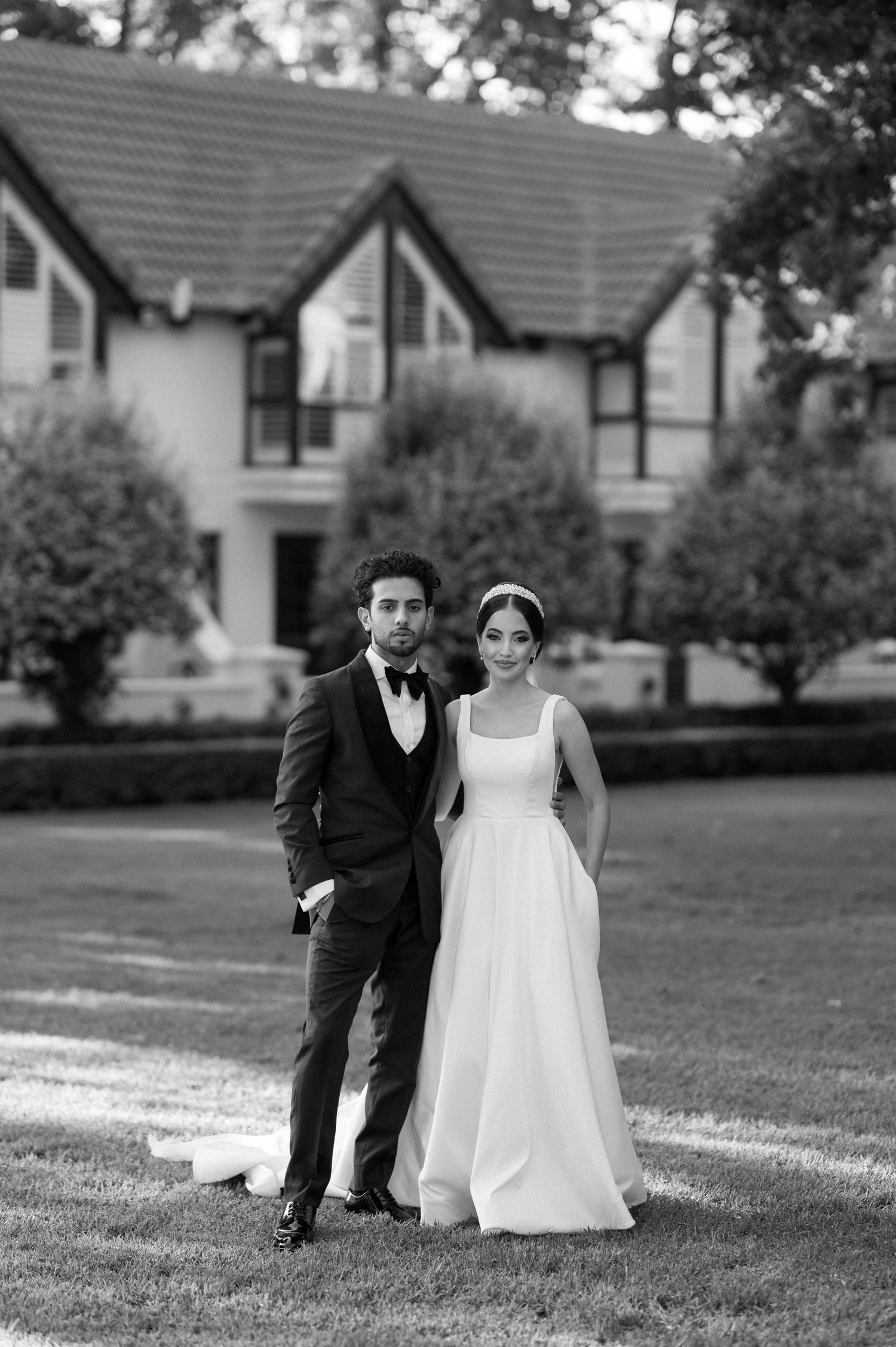 Black and white wedding portrait of couple standing in front of a Tudor-style house.