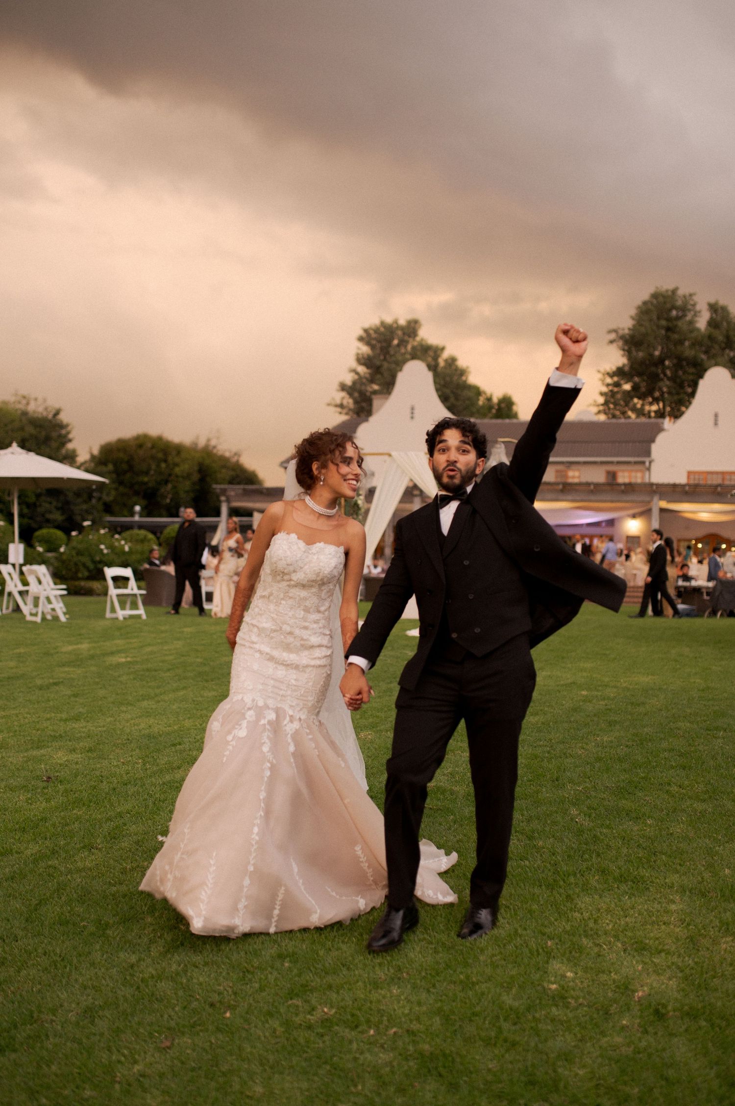 Wedding couple celebrating on lawn at sunset with dramatic sky backdrop.