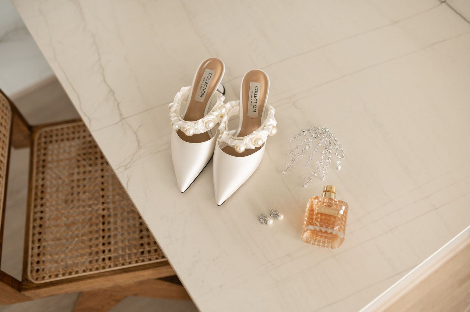 White designer mules and a perfume bottle arranged on marble in warm lighting.