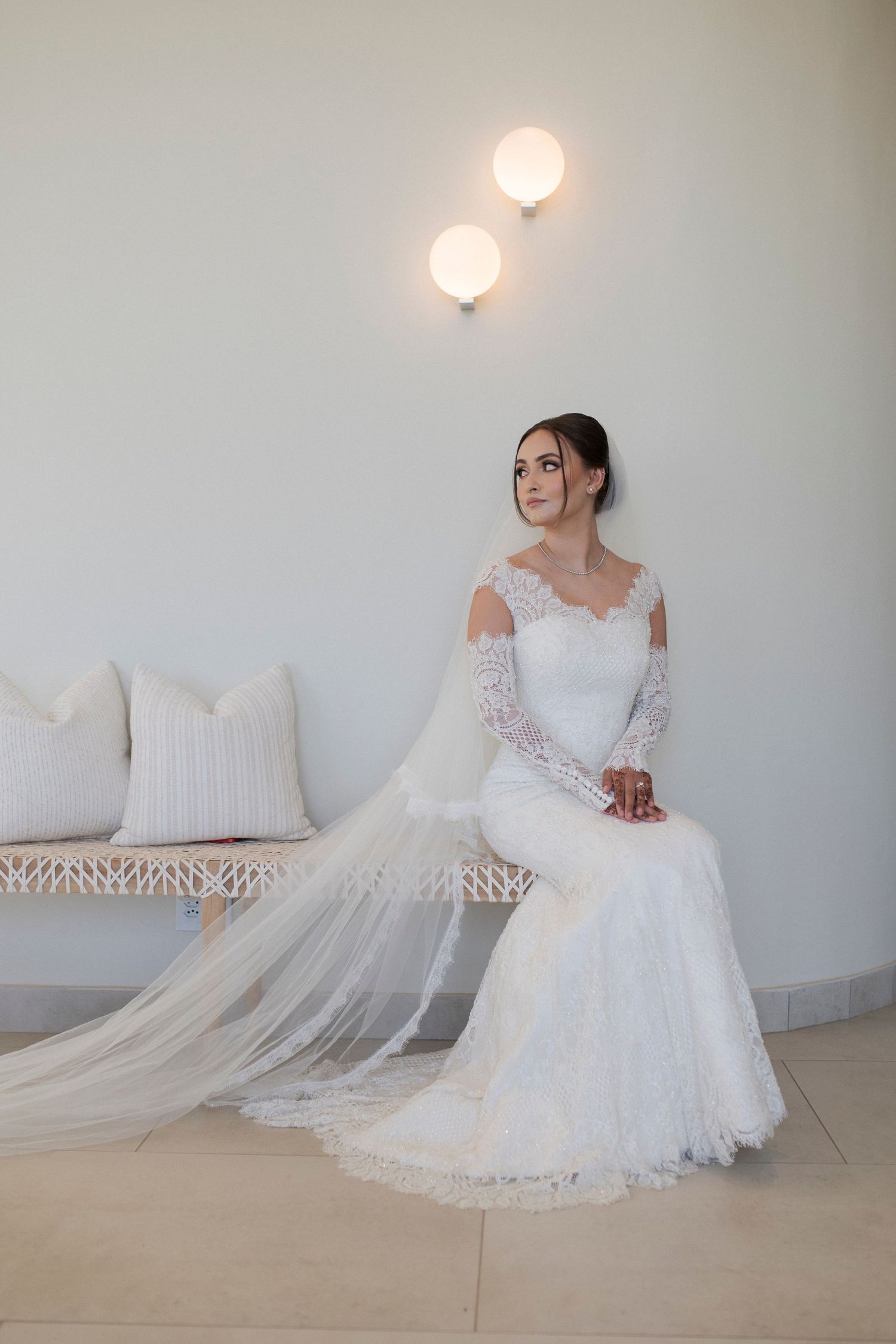 Bride in long-sleeve lace wedding gown sits elegantly on white bench under modern wall sconces.