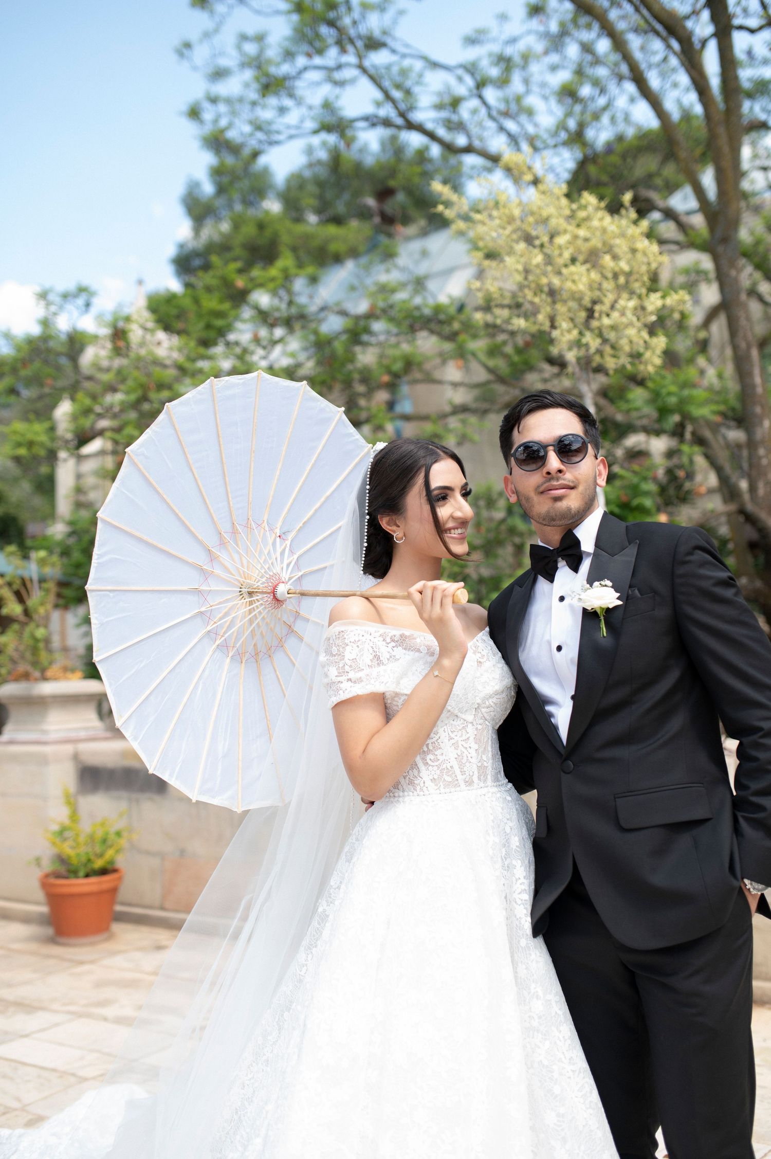 Happy couple with white parasol pose in garden courtyard, bride in lace gown and groom in tuxedo.