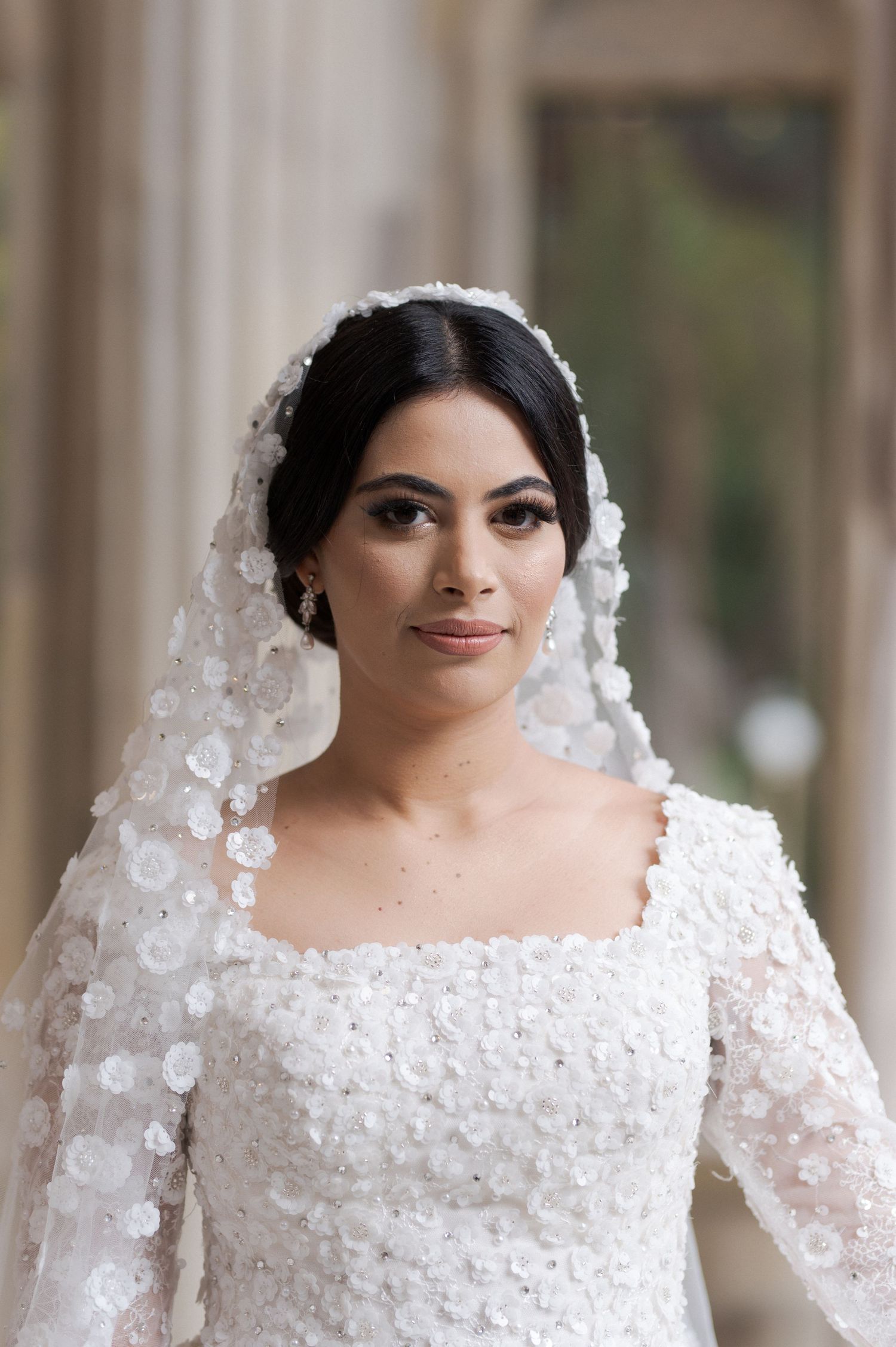 A bride wearing a long-sleeved lace wedding gown and veil poses for a formal portrait.