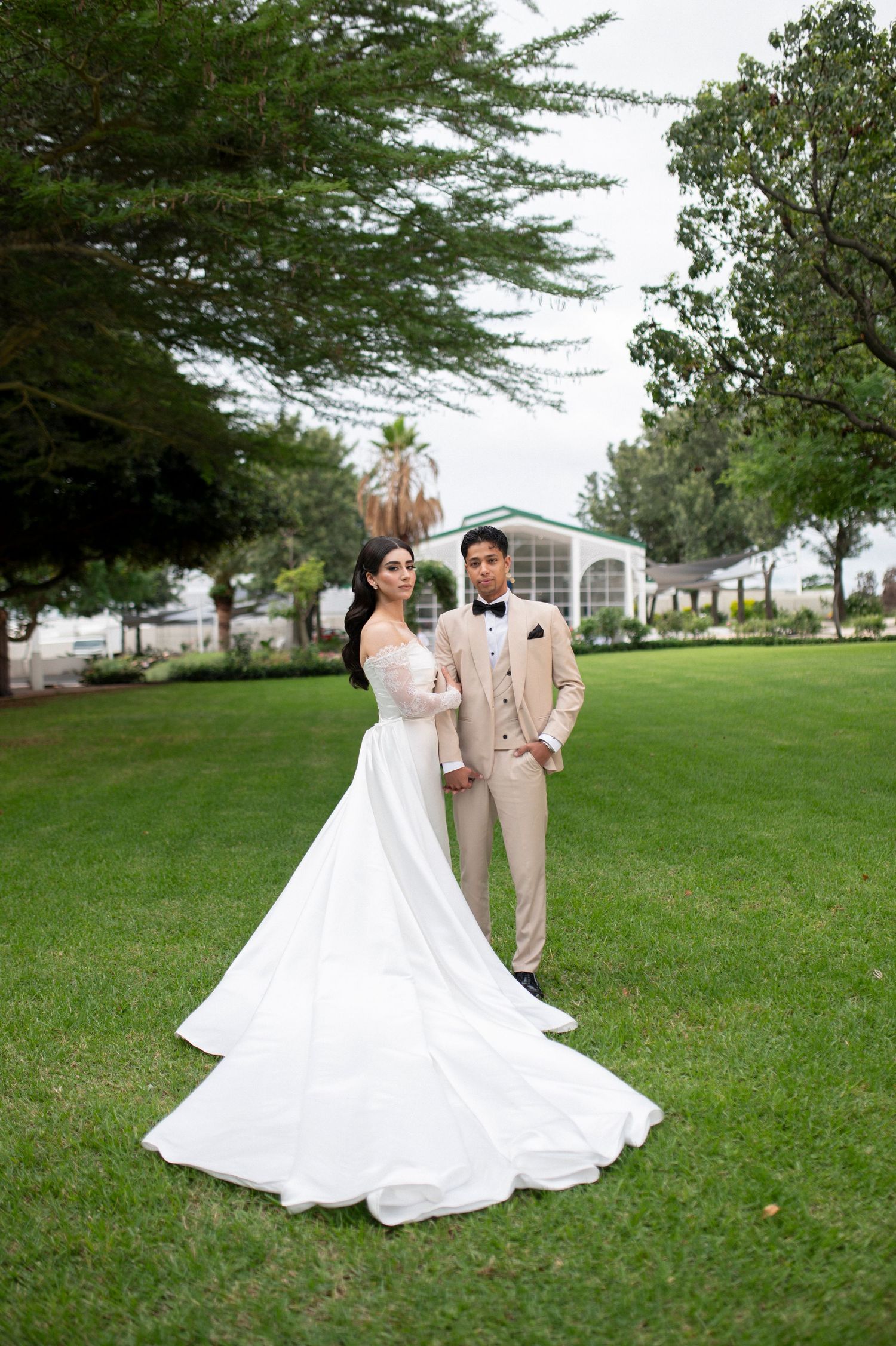Newlyweds pose on lawn with flowing white wedding gown and beige suit against church backdrop.