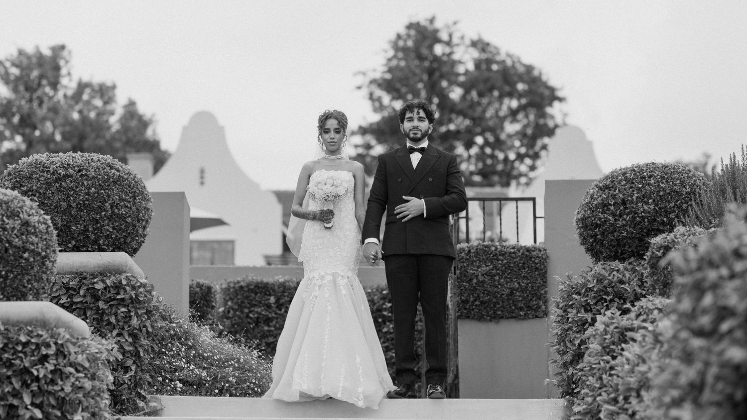 Black and white photo of couple walking down garden path with formal hedges.