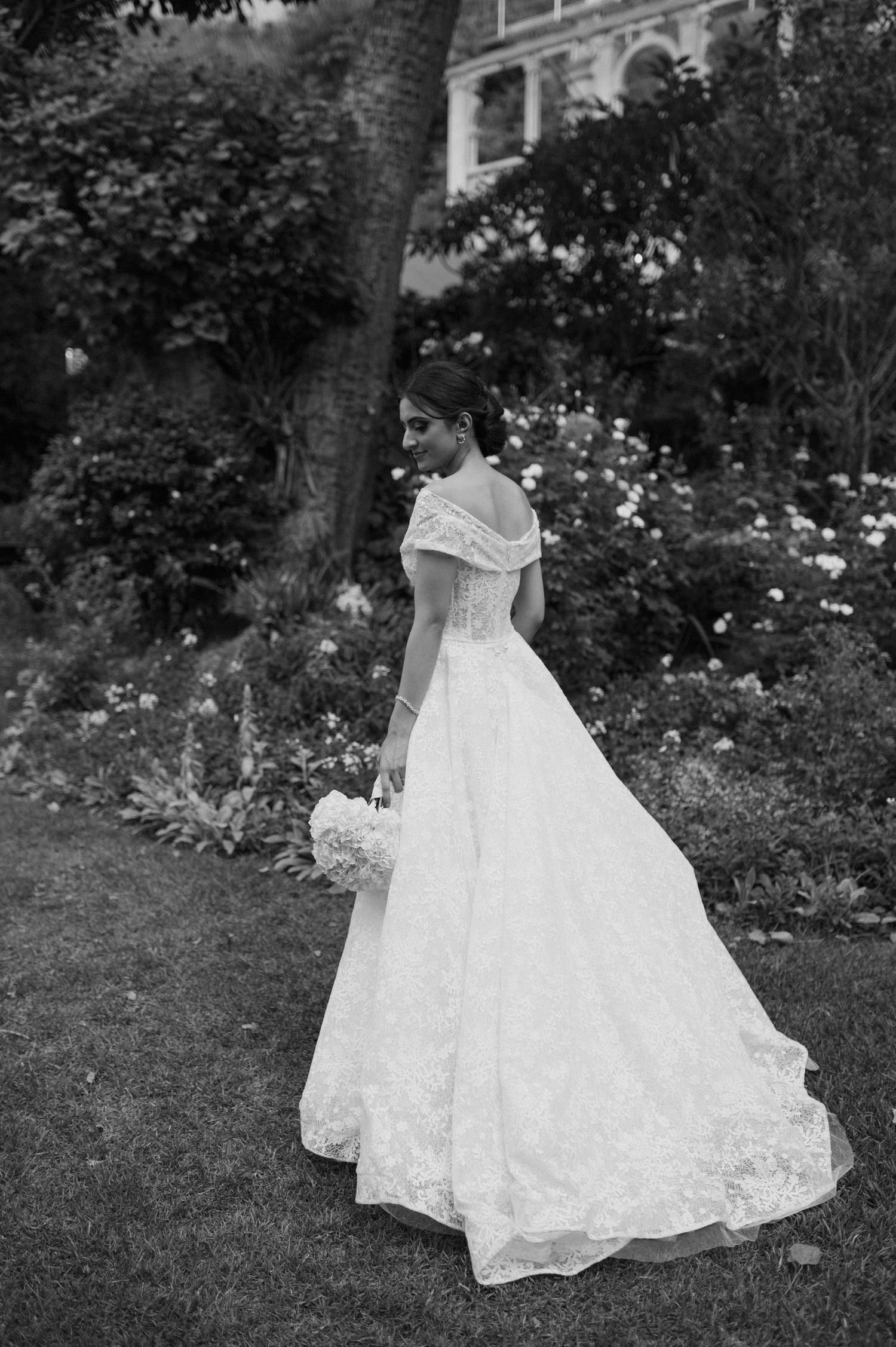 Black and white portrait of a bride in a classic off-shoulder gown holding a bouquet in a garden.