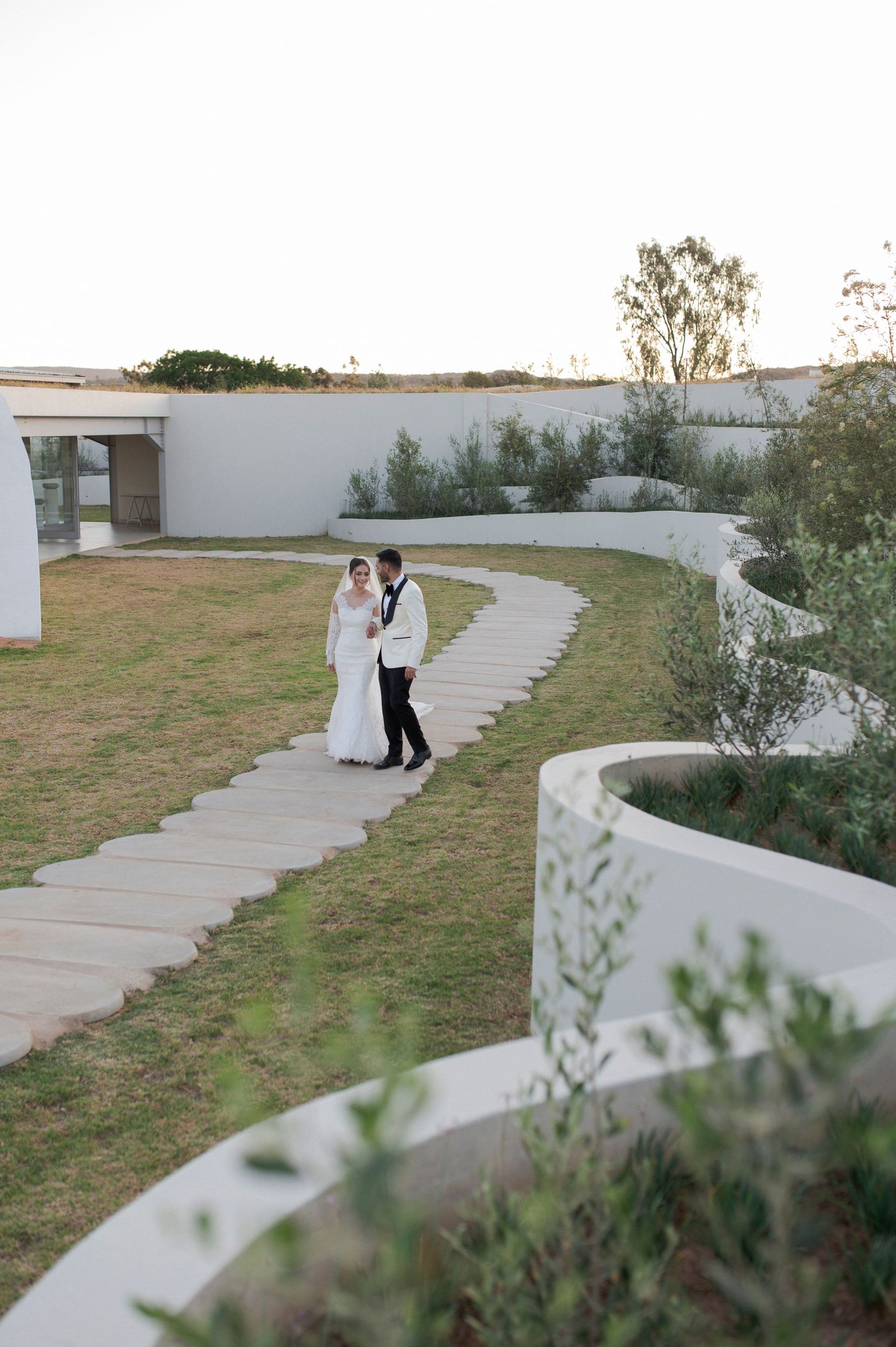 A couple walks along a curved garden path surrounded by white walls and lush greenery.