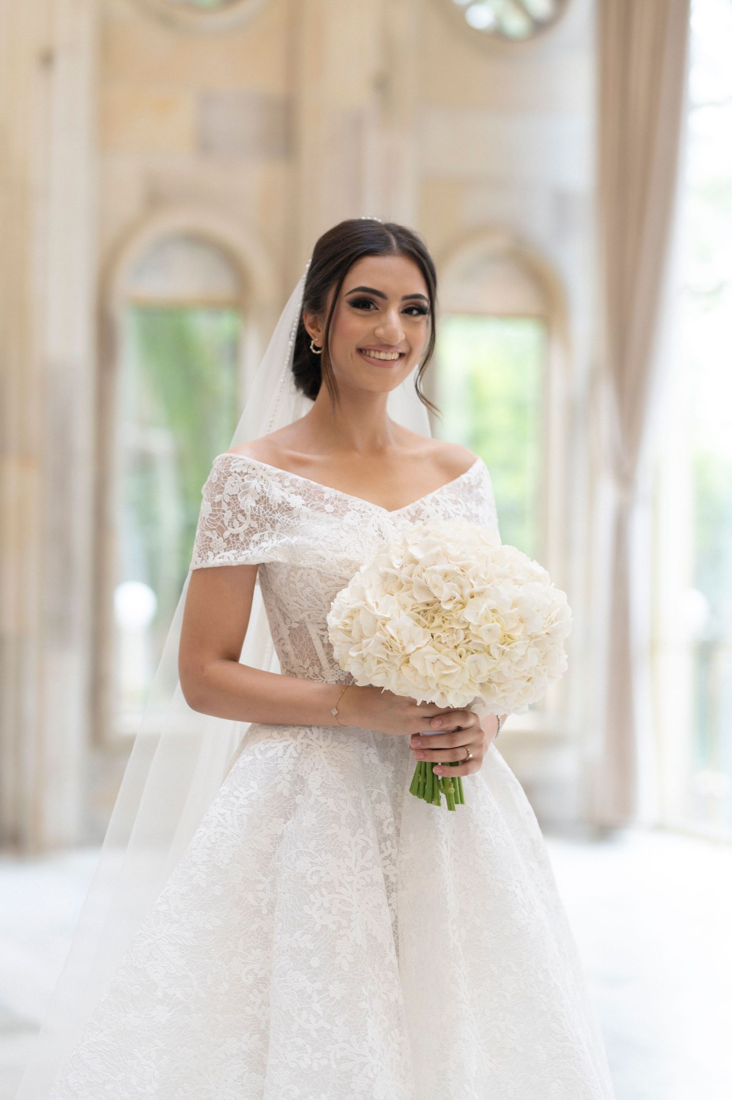 An elegant bride in a white lace gown holding a luxurious white bouquet in a sunlit room.
