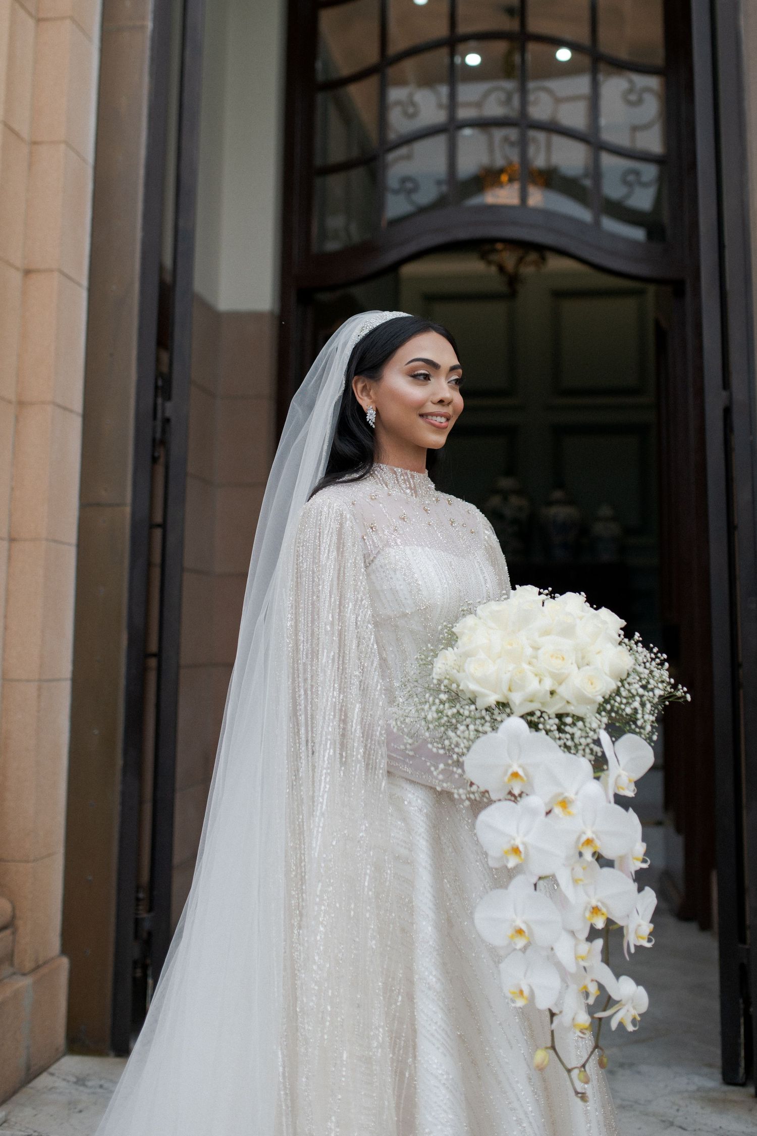 A bride in a sparkly white gown holds a stunning cascading bouquet featuring white orchids and roses.