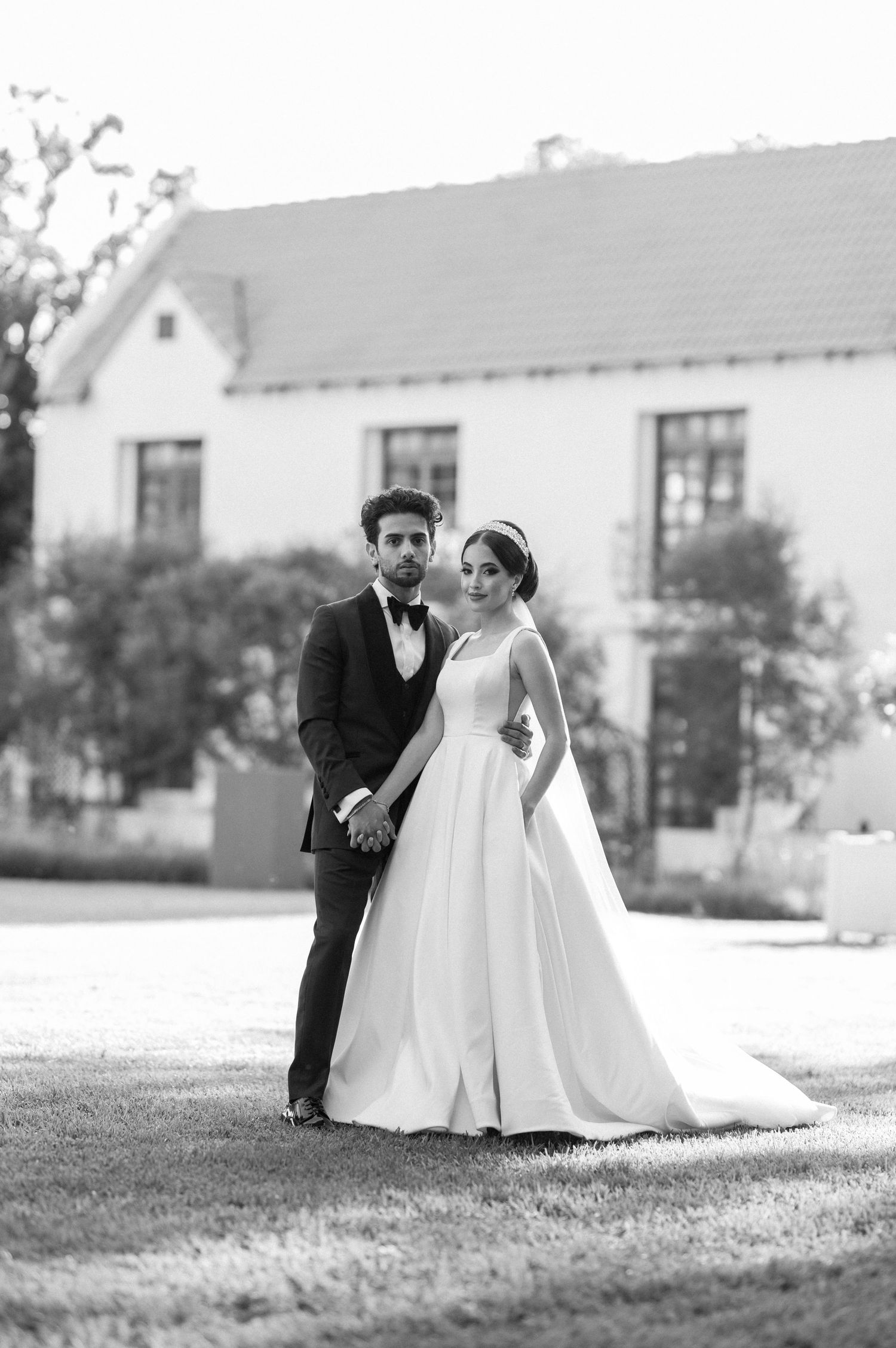 Black and white portrait of newlyweds standing together on grass with house in background.