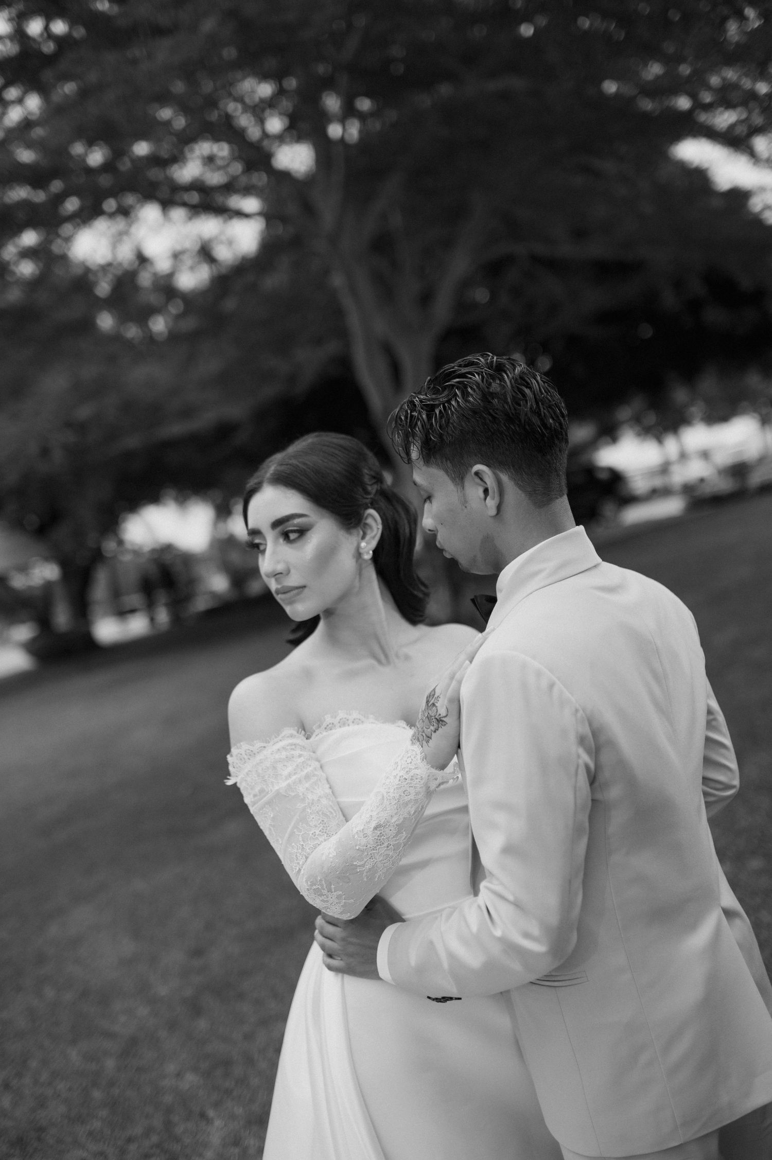 Elegant bride in off-shoulder lace gown embraces groom in white suit during outdoor wedding photoshoot.