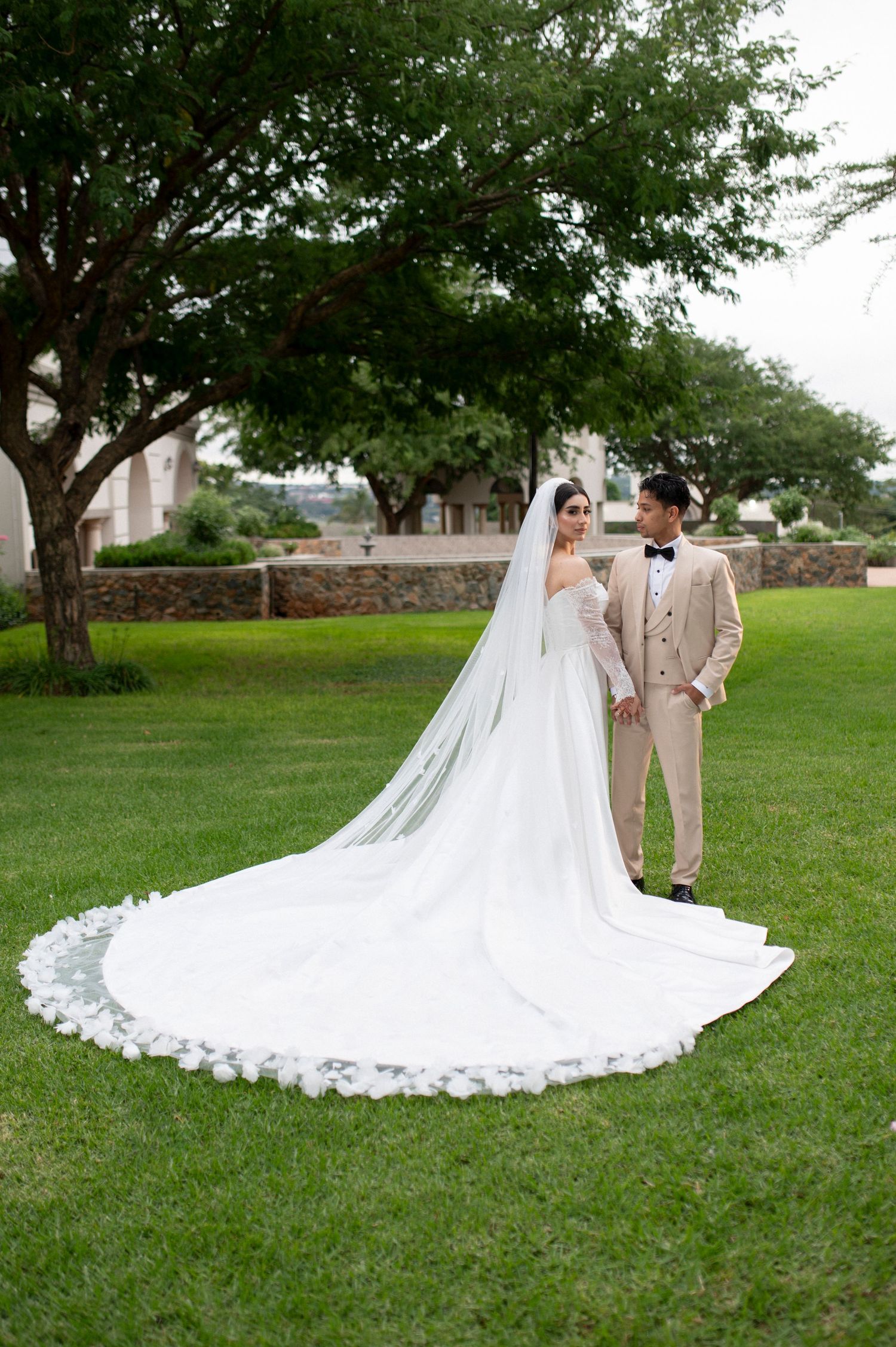 A wedding couple poses on grass with bride's cathedral-length lace veil spread beautifully.
