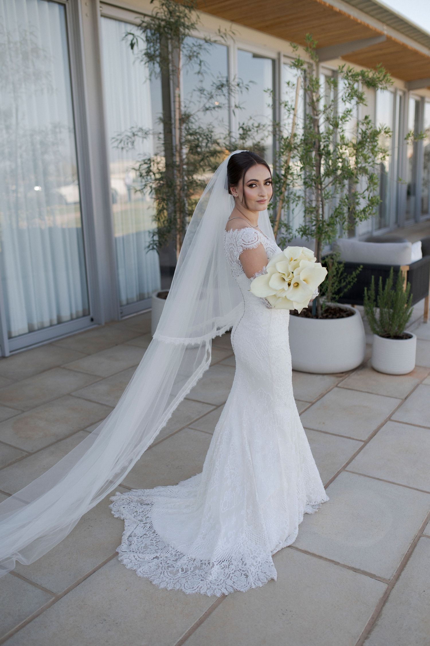 Bride in form-fitting lace gown with cathedral veil poses on outdoor terrace with white bouquet.