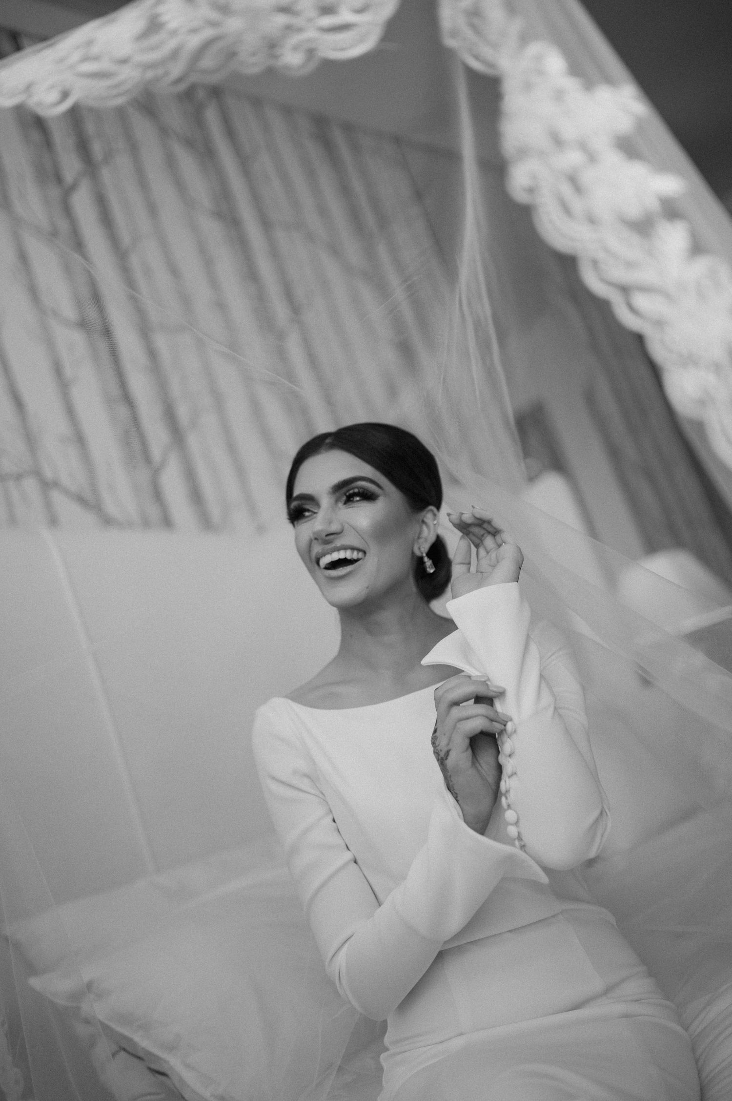 Black and white portrait of a joyful bride in a long-sleeved wedding dress sitting under a veil.