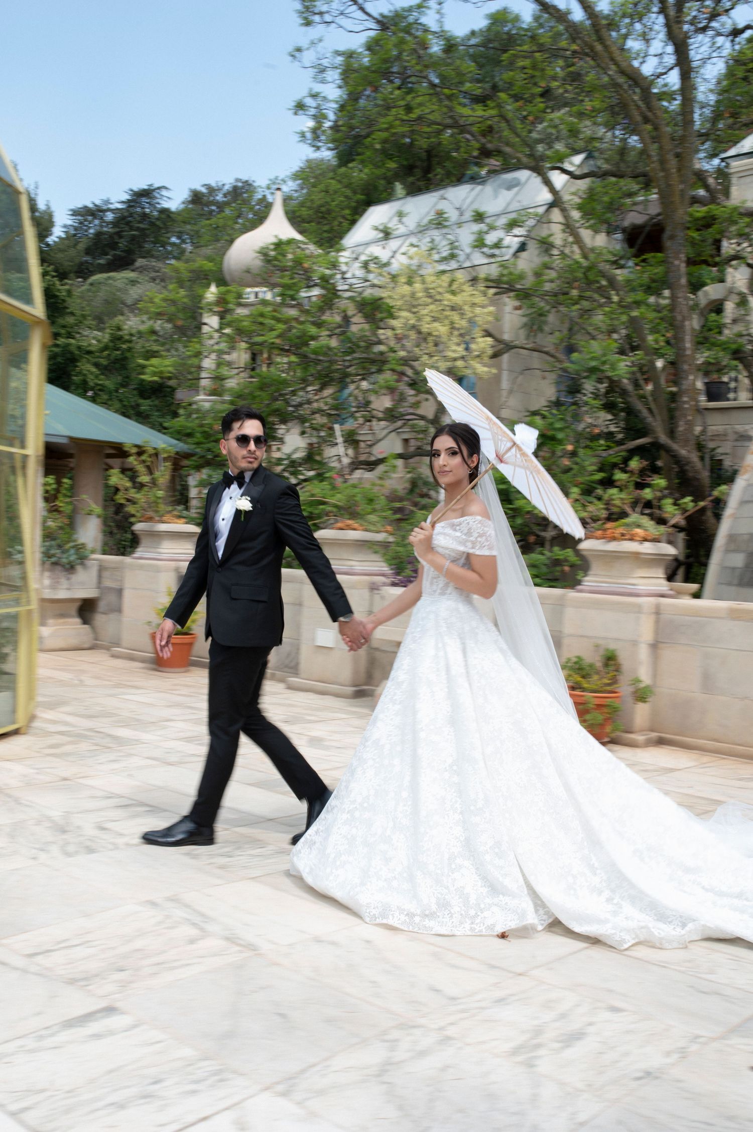 Newlyweds holding hands while walking with parasol through garden courtyard.