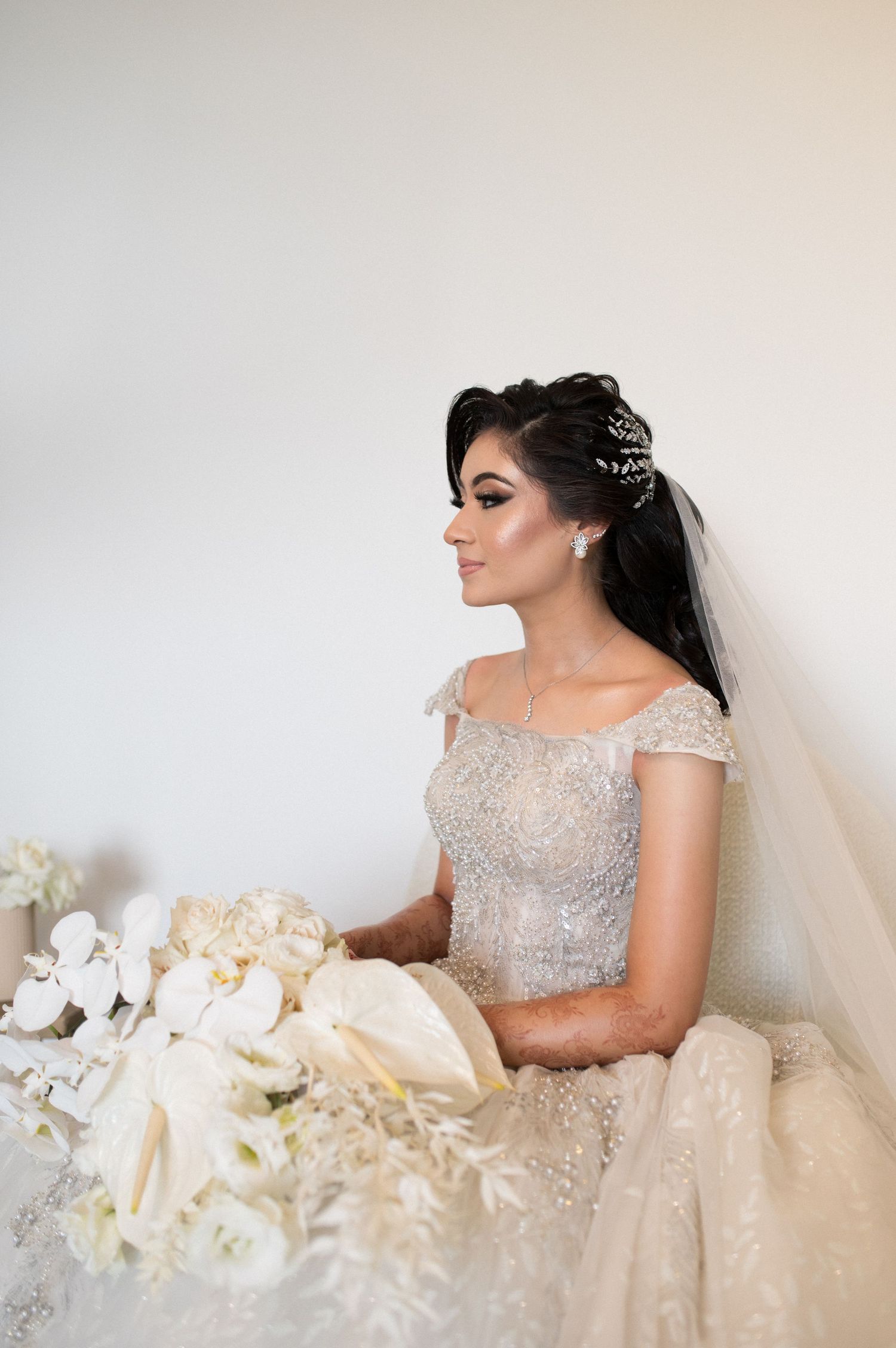 Bride in embellished gown holding white bouquet against neutral background.