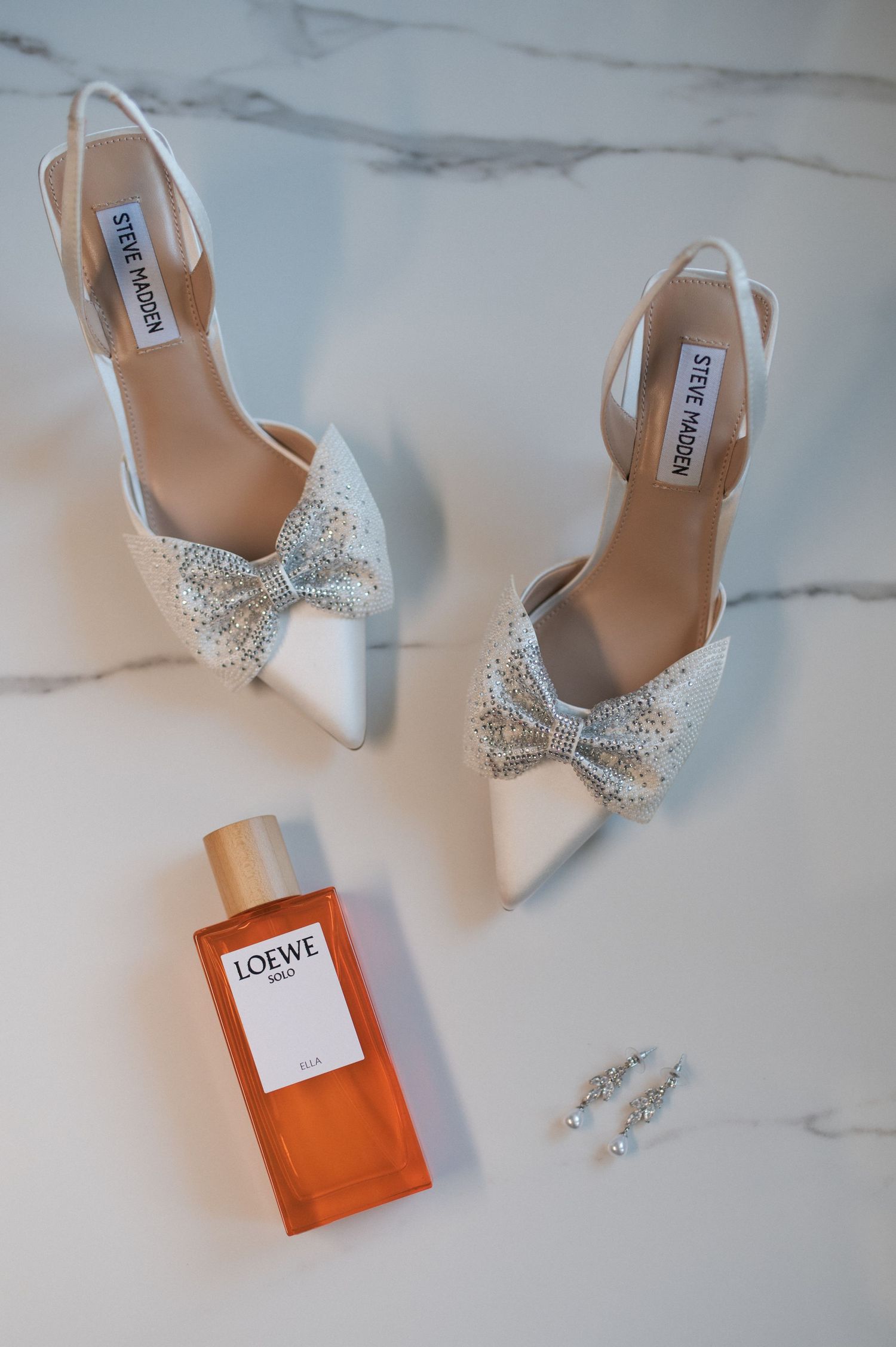 White bridal shoes with crystal bows displayed with Loewe perfume and jewelry on marble surface.