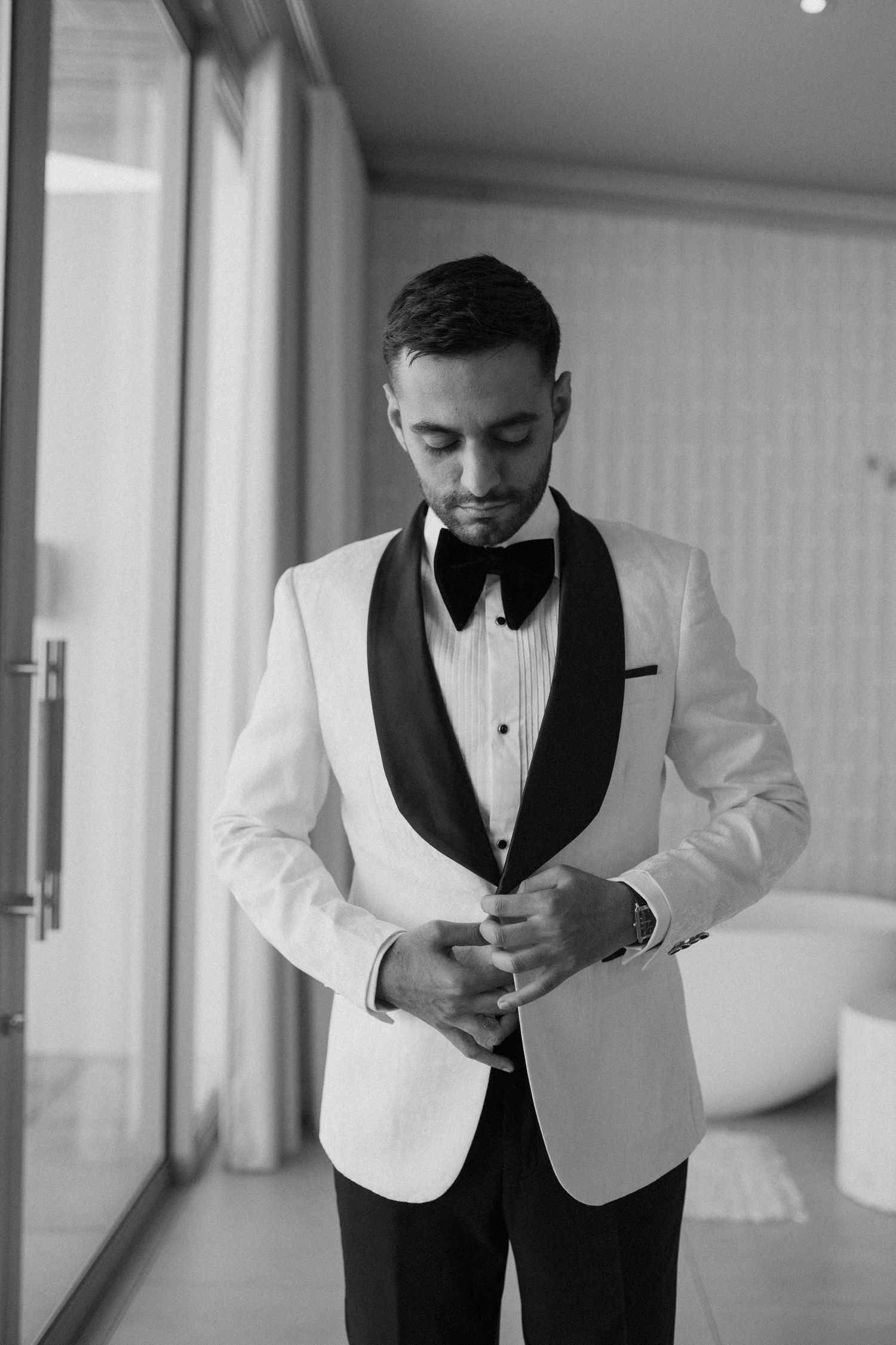 Black and white portrait of groom adjusting cufflinks in white dinner jacket.