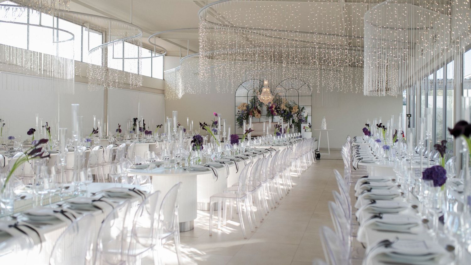 Modern all-white wedding reception venue with crystal chandeliers and ghost chairs.