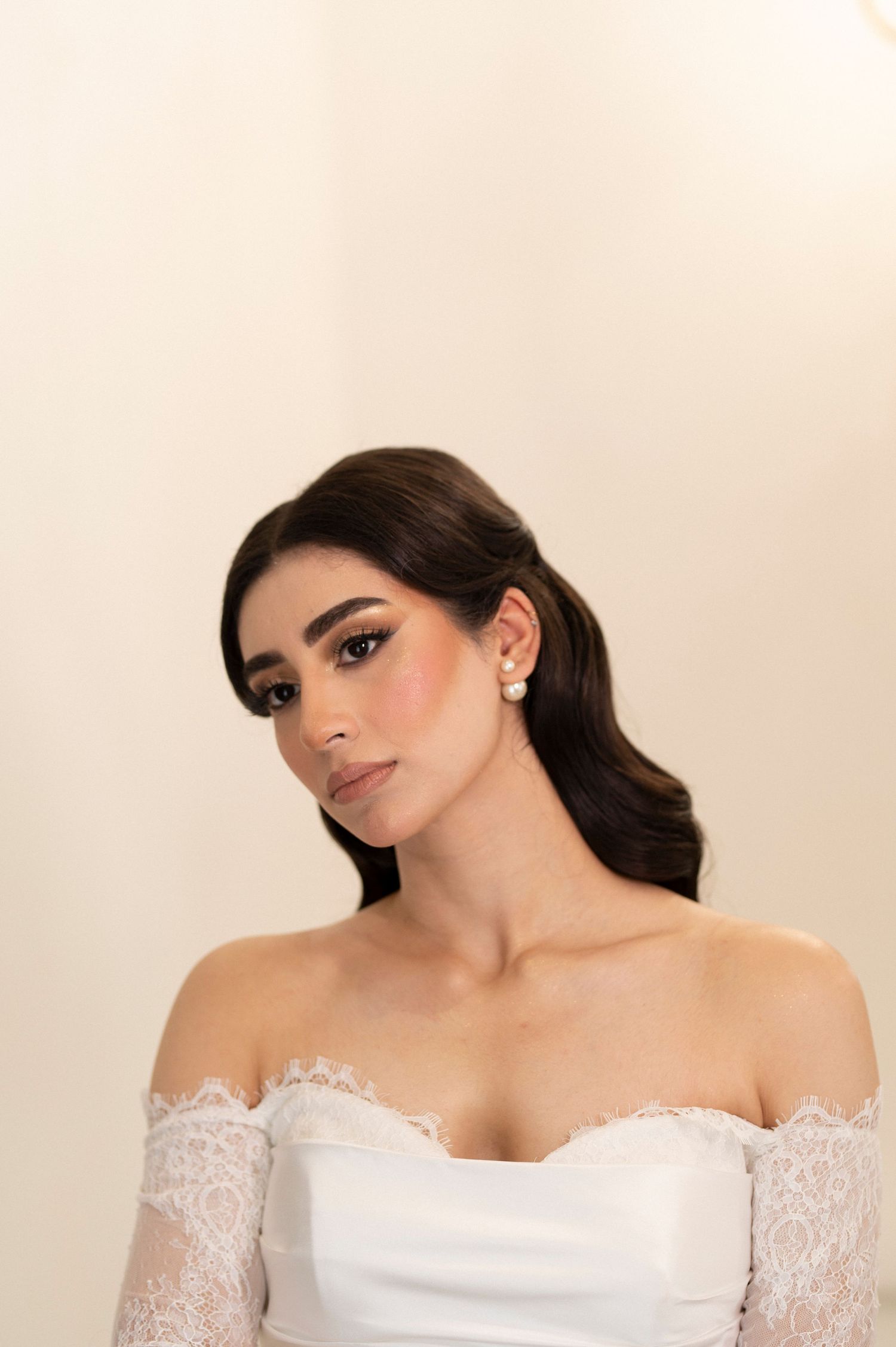 A bride in an elegant off-shoulder lace wedding dress poses for a portrait with soft makeup and dark wavy hair.