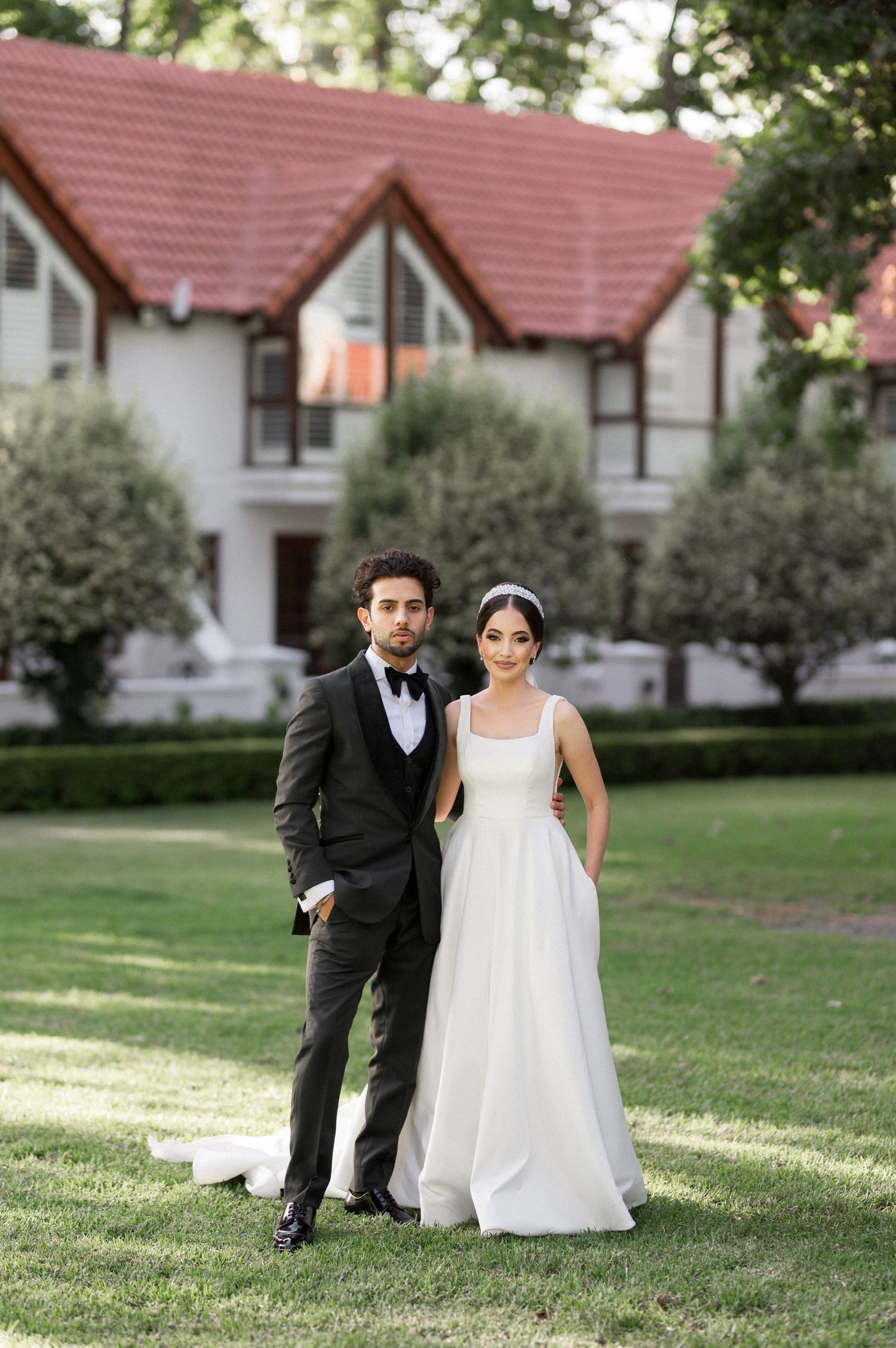 Wedding couple poses on lawn in front of white house with red roof.