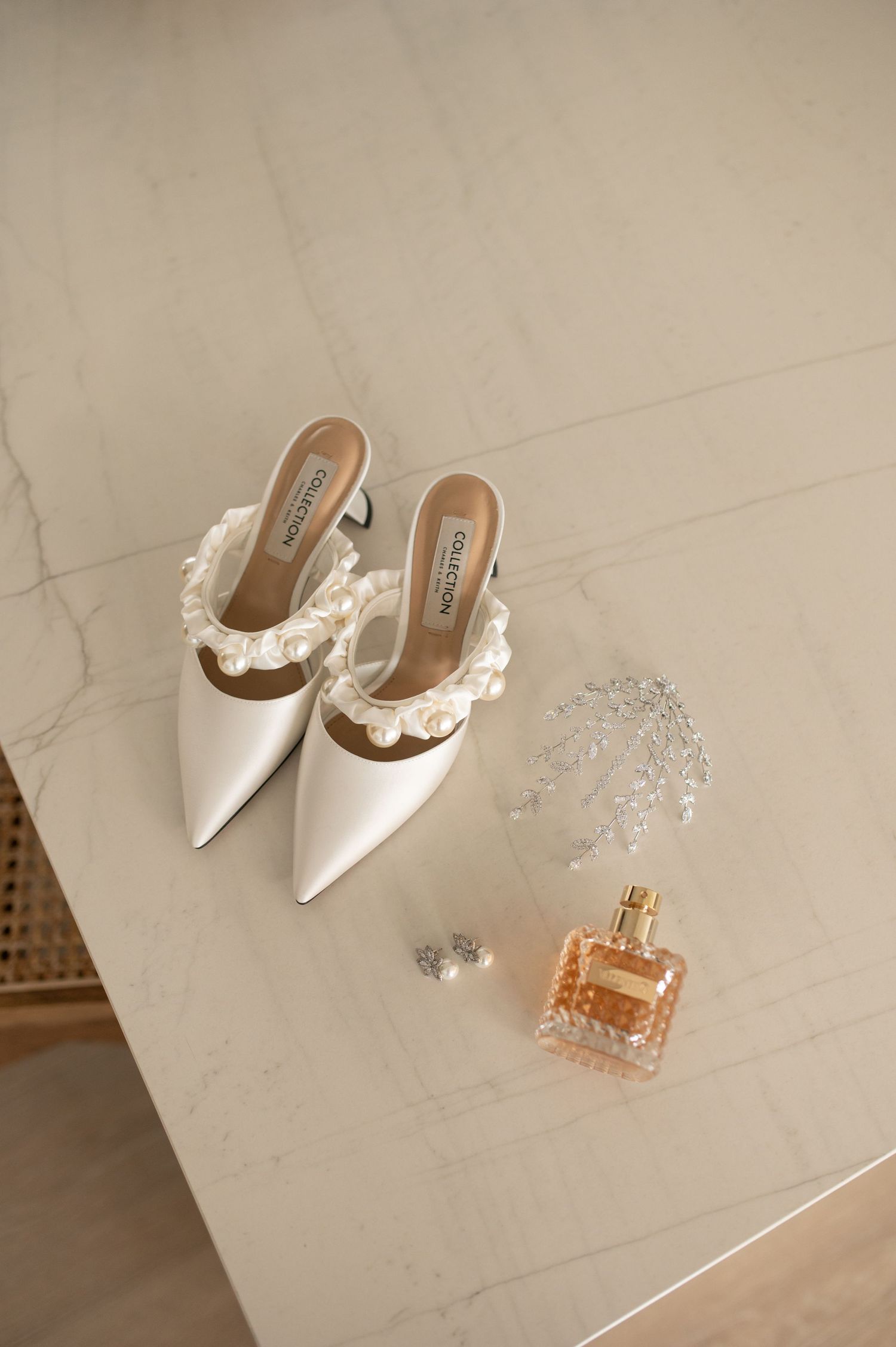 White pointed-toe bridal shoes with pearl embellishments displayed with perfume and accessories.