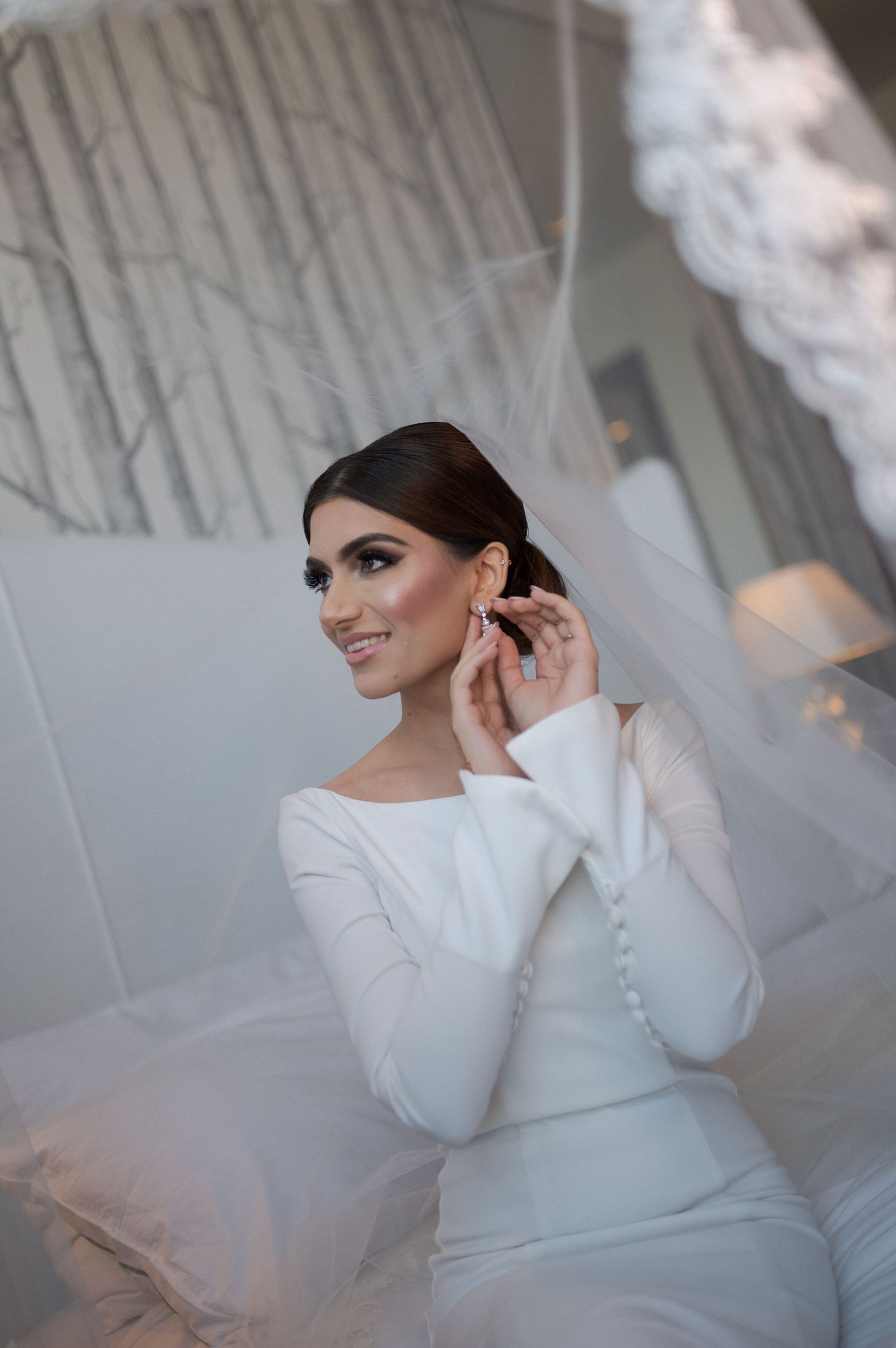 Bride in sleek white long-sleeved wedding dress adjusting earring in preparation room.