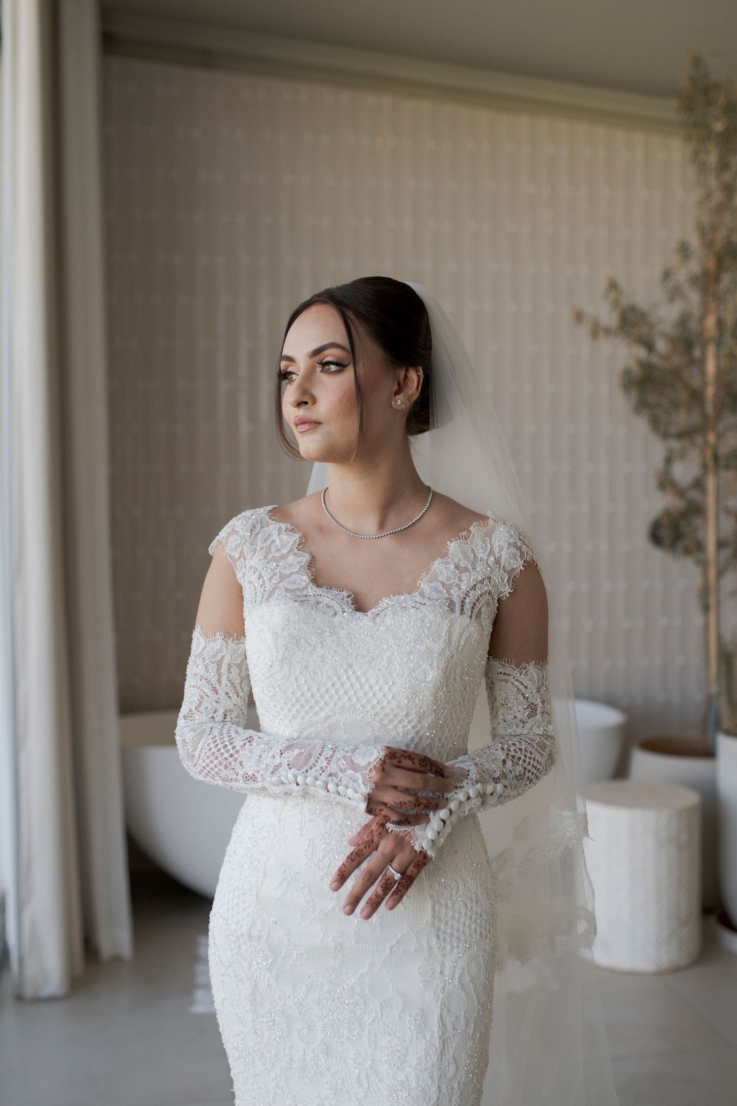 Bride poses in fitted lace wedding dress with sheer long sleeves against white interior.