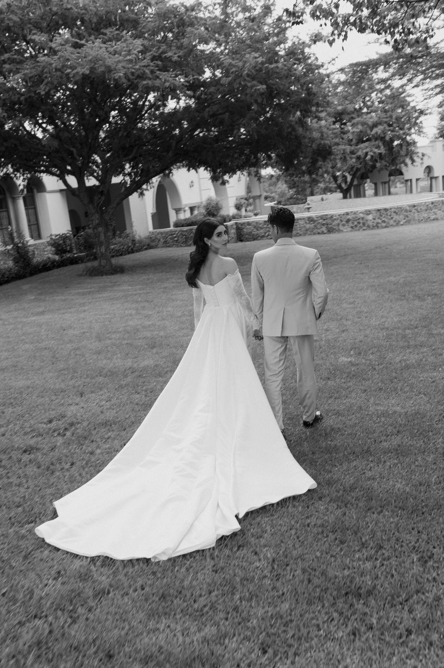 A couple walking on grass with bride's long white dress train flowing behind them.