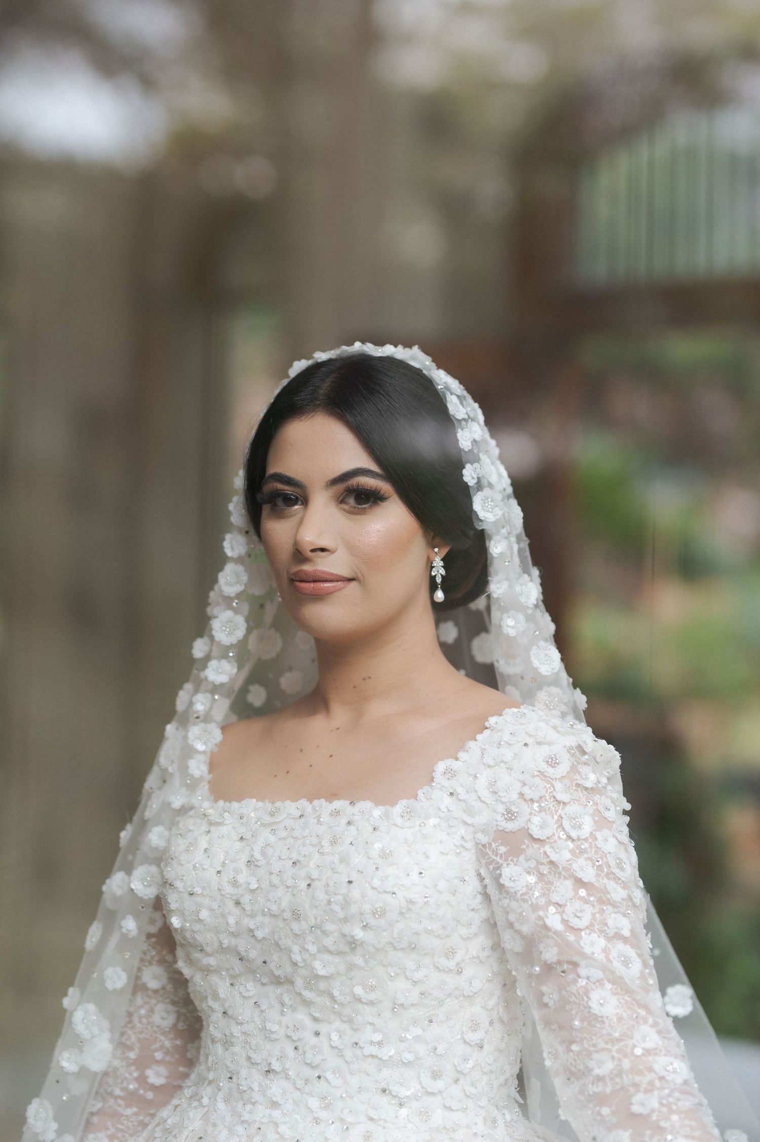 A bride in an ornate white lace dress and floral veil poses for a wedding portrait.
