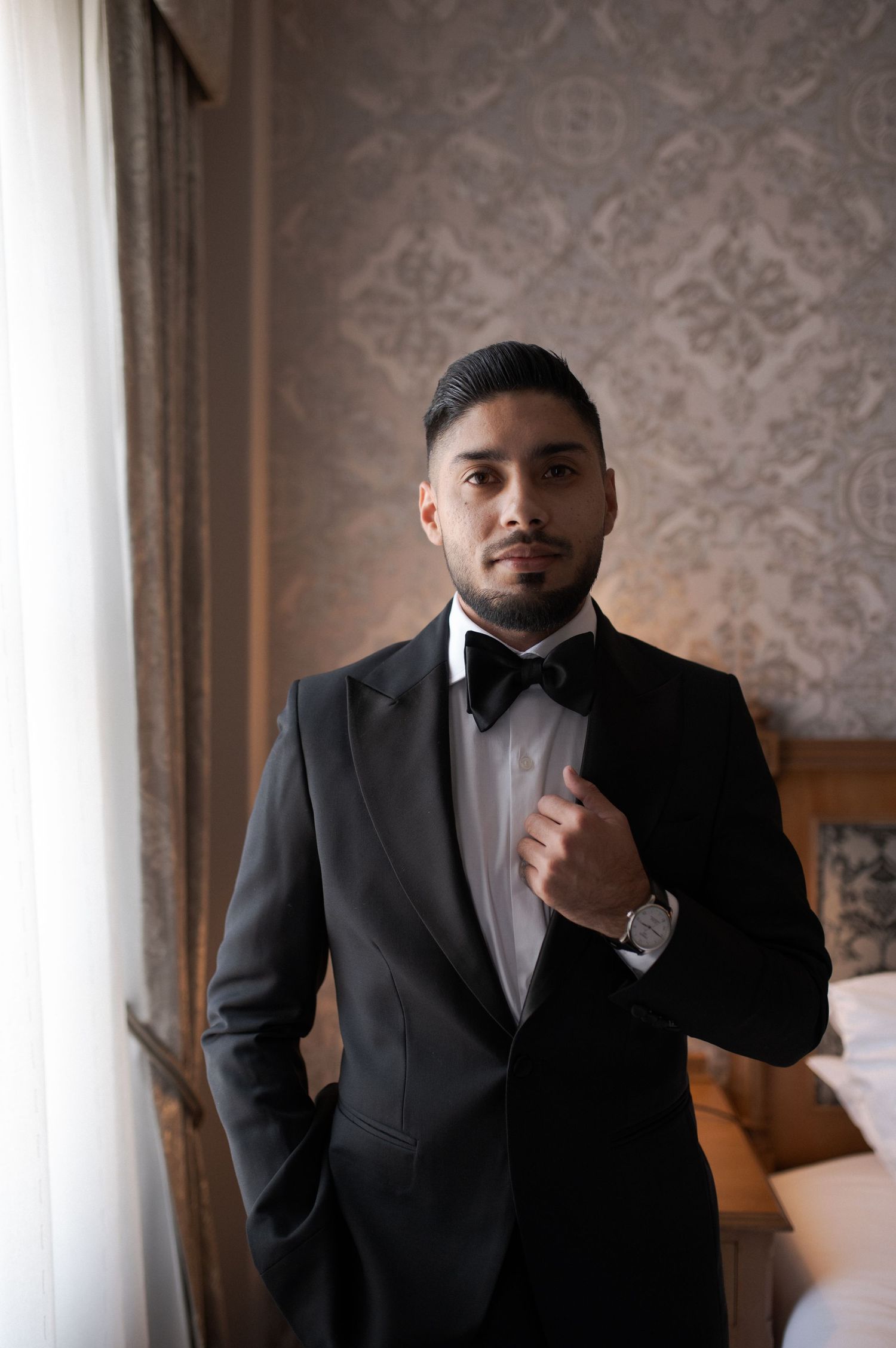 Well-dressed man in black tuxedo and bowtie poses by window.