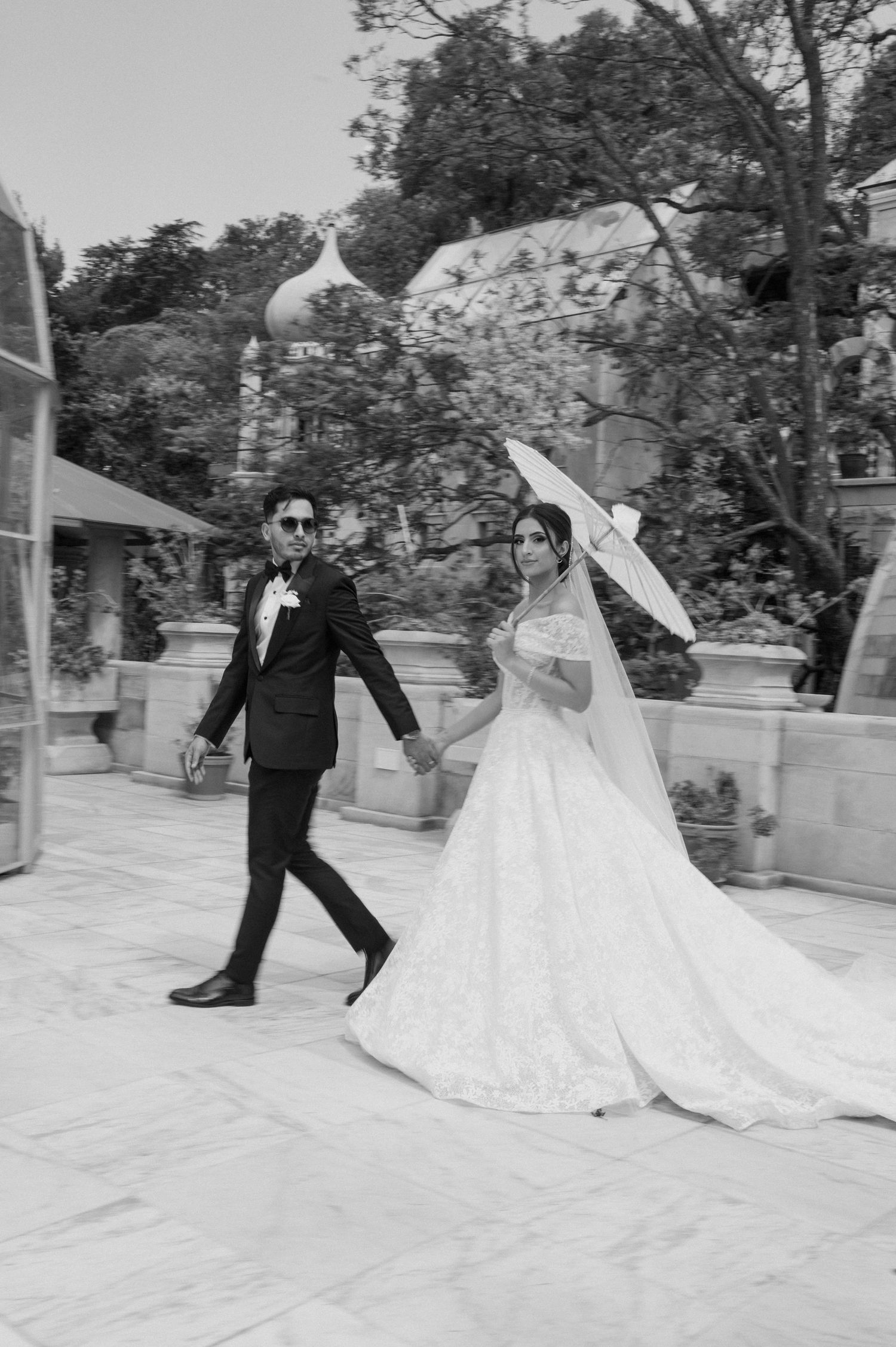 Black and white photo of bride and groom walking with parasol in elegant outdoor setting.