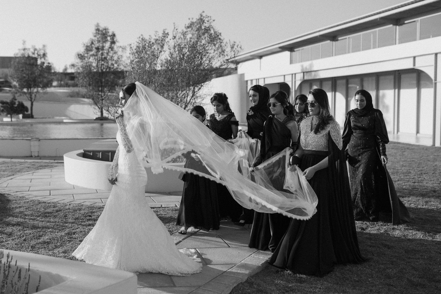 Bridesmaids in black dresses help arrange bride's long flowing veil outdoors.