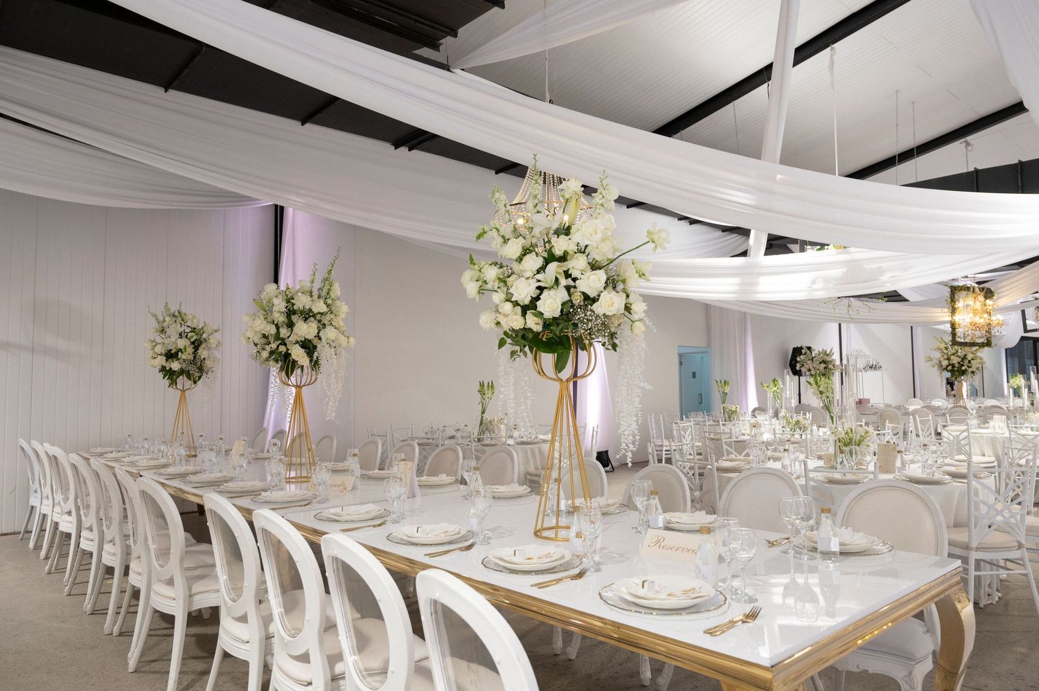 Elegant white wedding reception setup with long tables, gold accents, and floral centerpieces.