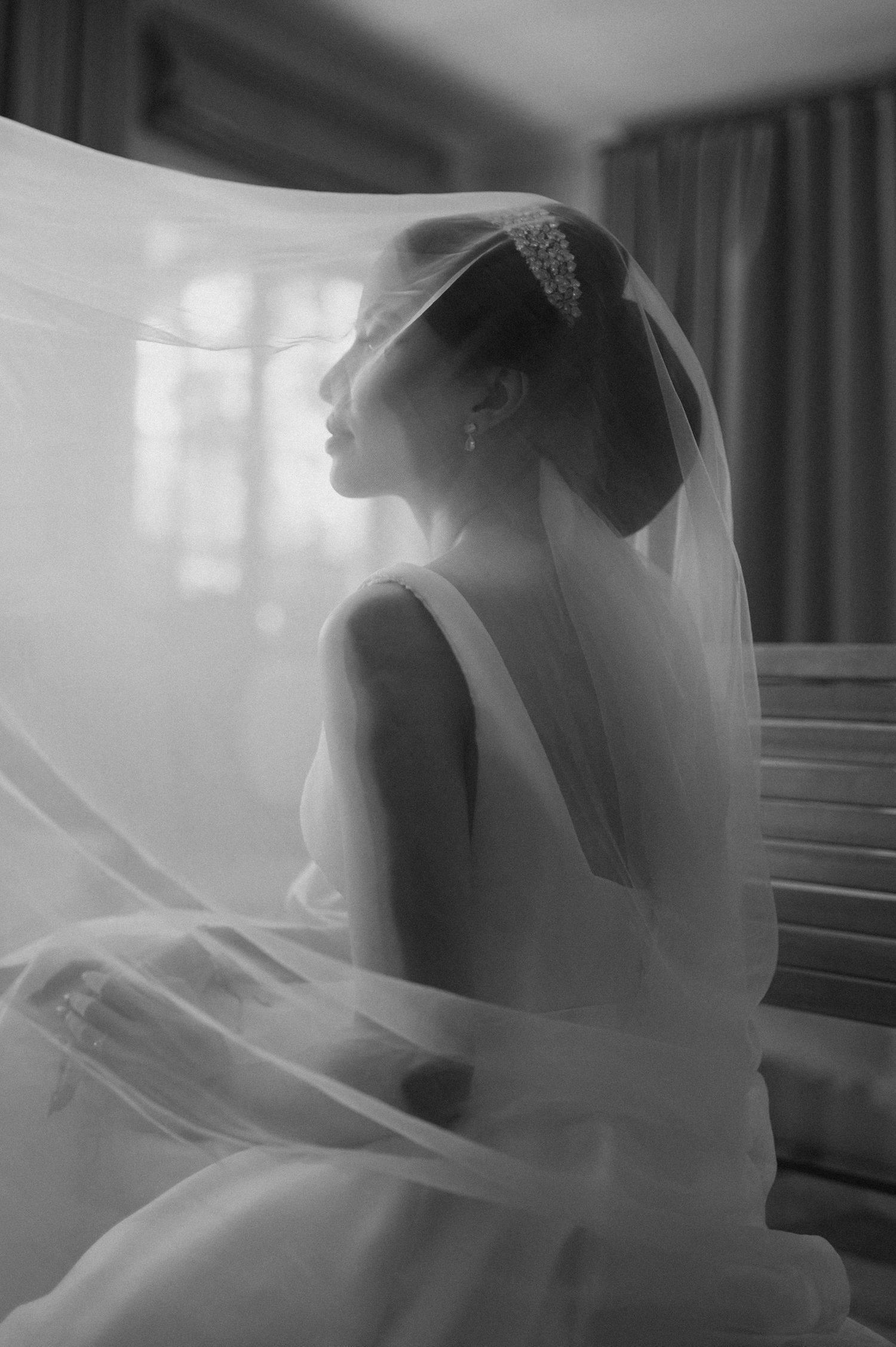 Black and white silhouette of bride in wedding dress and veil sitting in sunlit room.