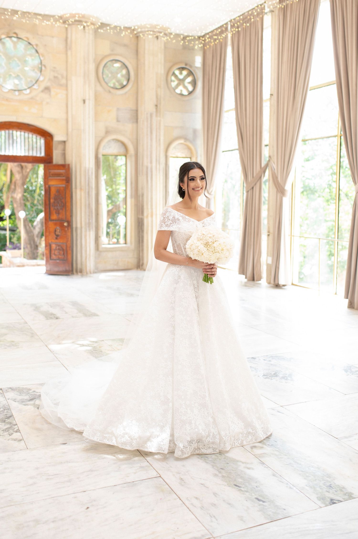 Bride wearing classic white ballgown poses with bouquet in sunlit room with ornate architectural details.