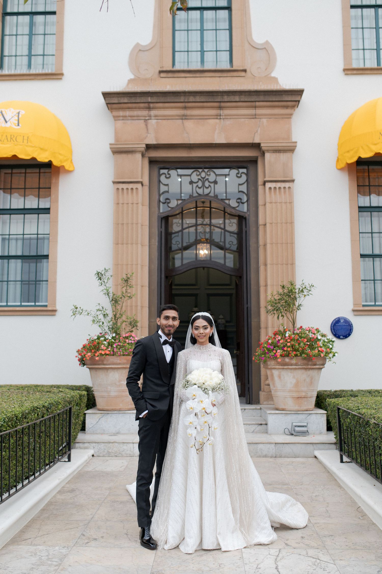 A newlywed couple stands on steps of a building with yellow awnings and decorative entrance.