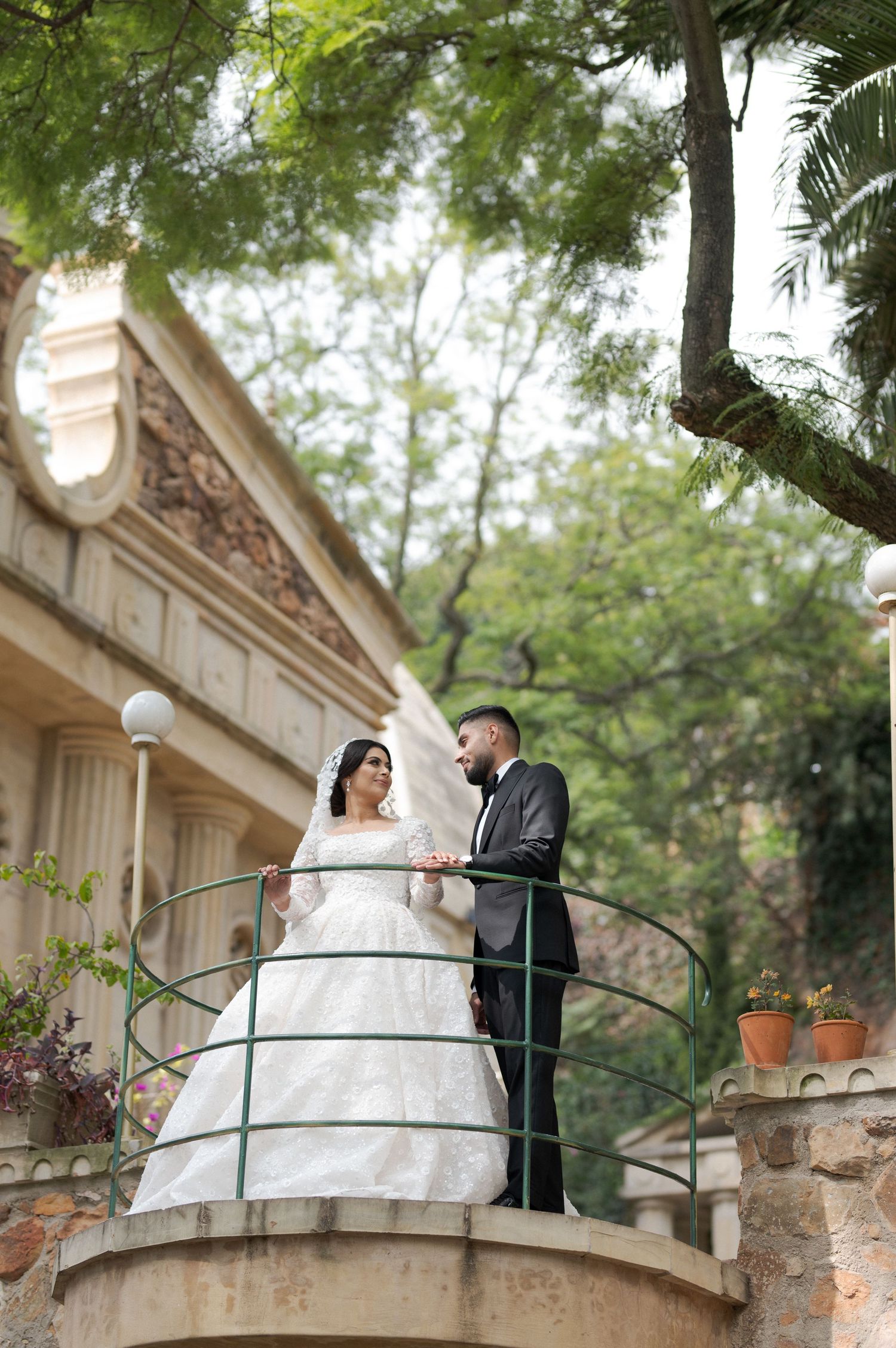 Newlyweds share a romantic moment on an ornate balcony surrounded by lush greenery.