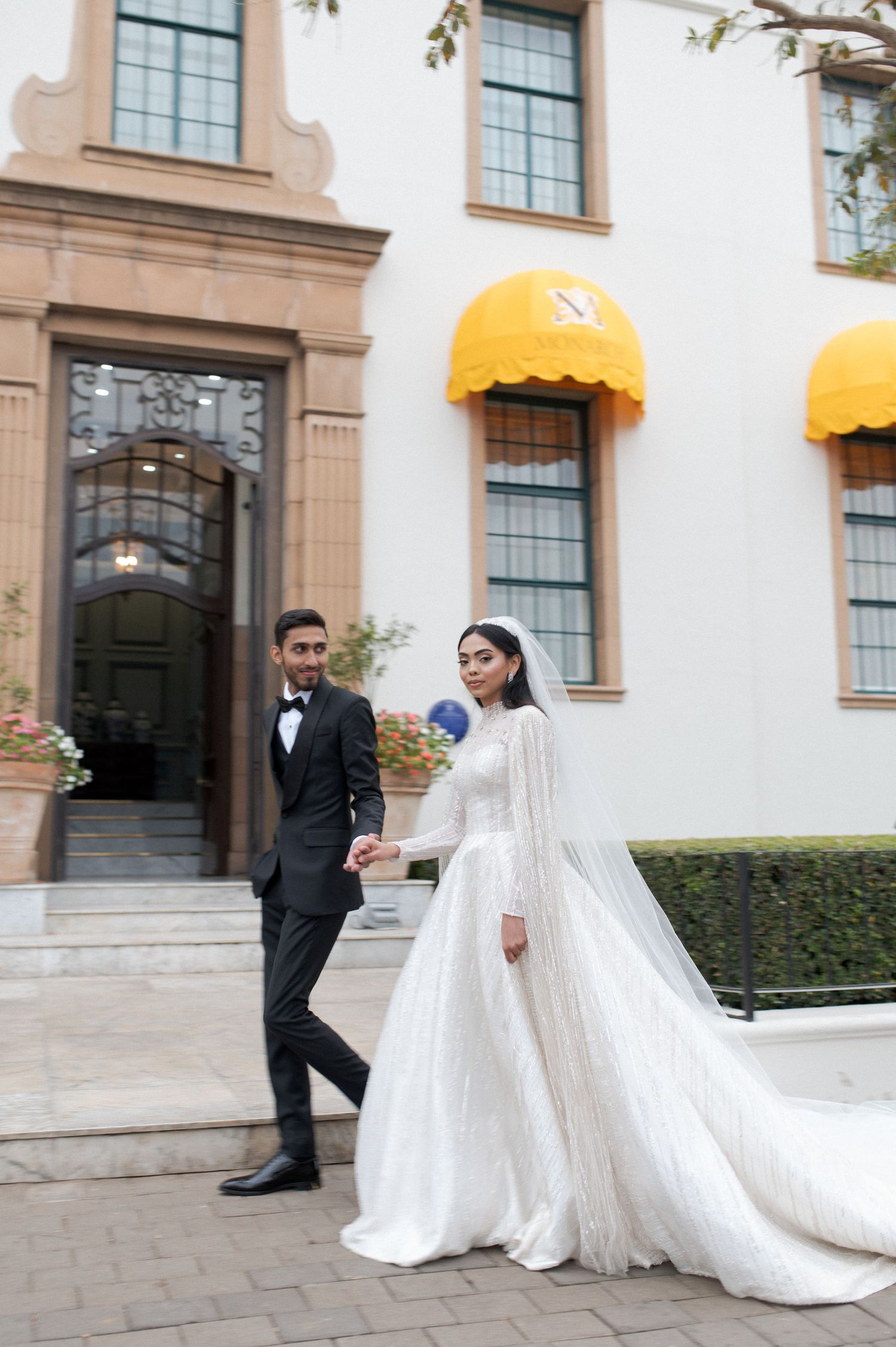 A wedding couple stands outside a building with yellow awnings and tall windows in an elegant setting.