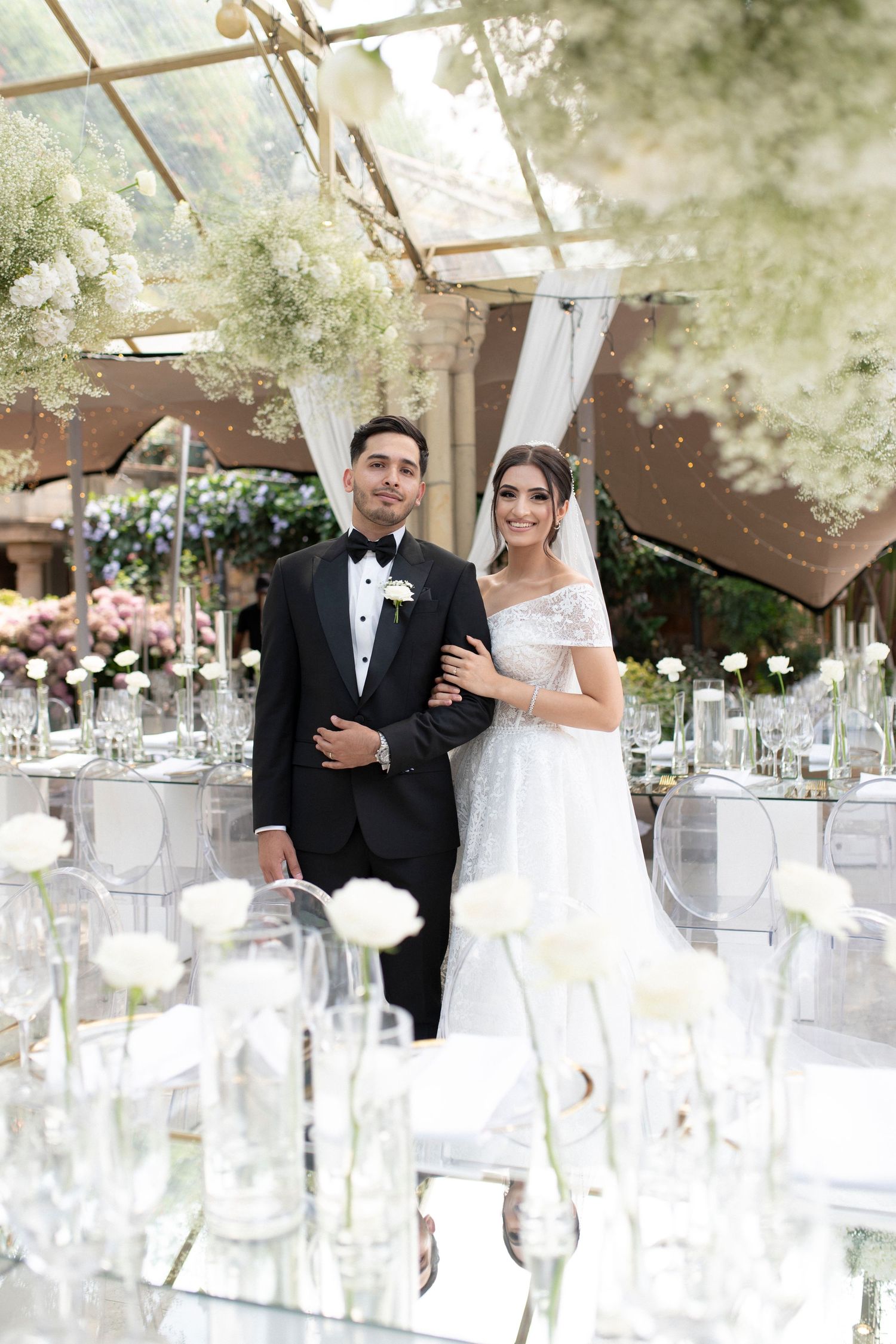 An elegant wedding reception setting with glass tables, white flowers, and a couple posed among crystal decor.
