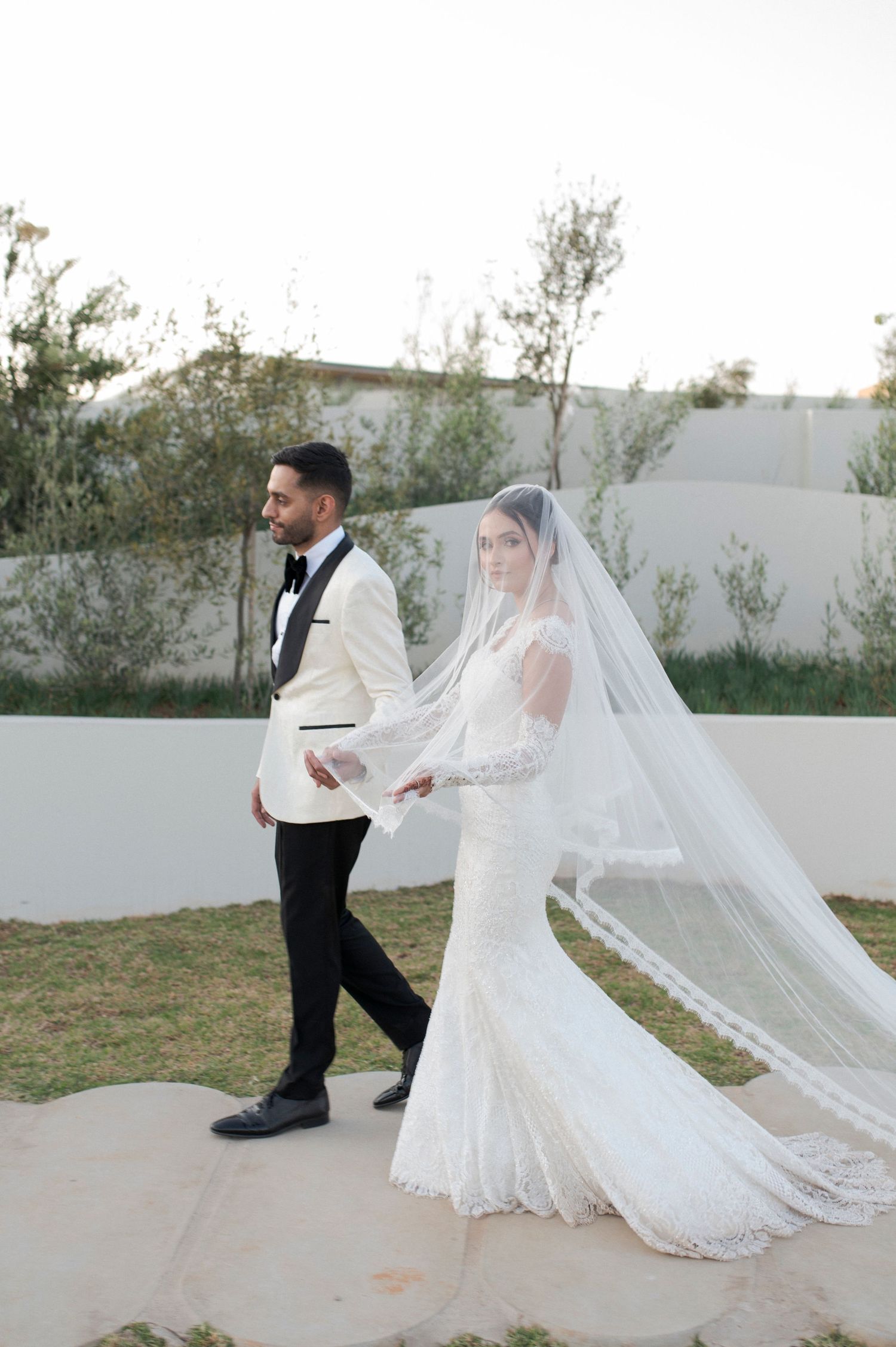 A couple walks hand in hand on a garden path with a dramatic flowing veil trailing behind.