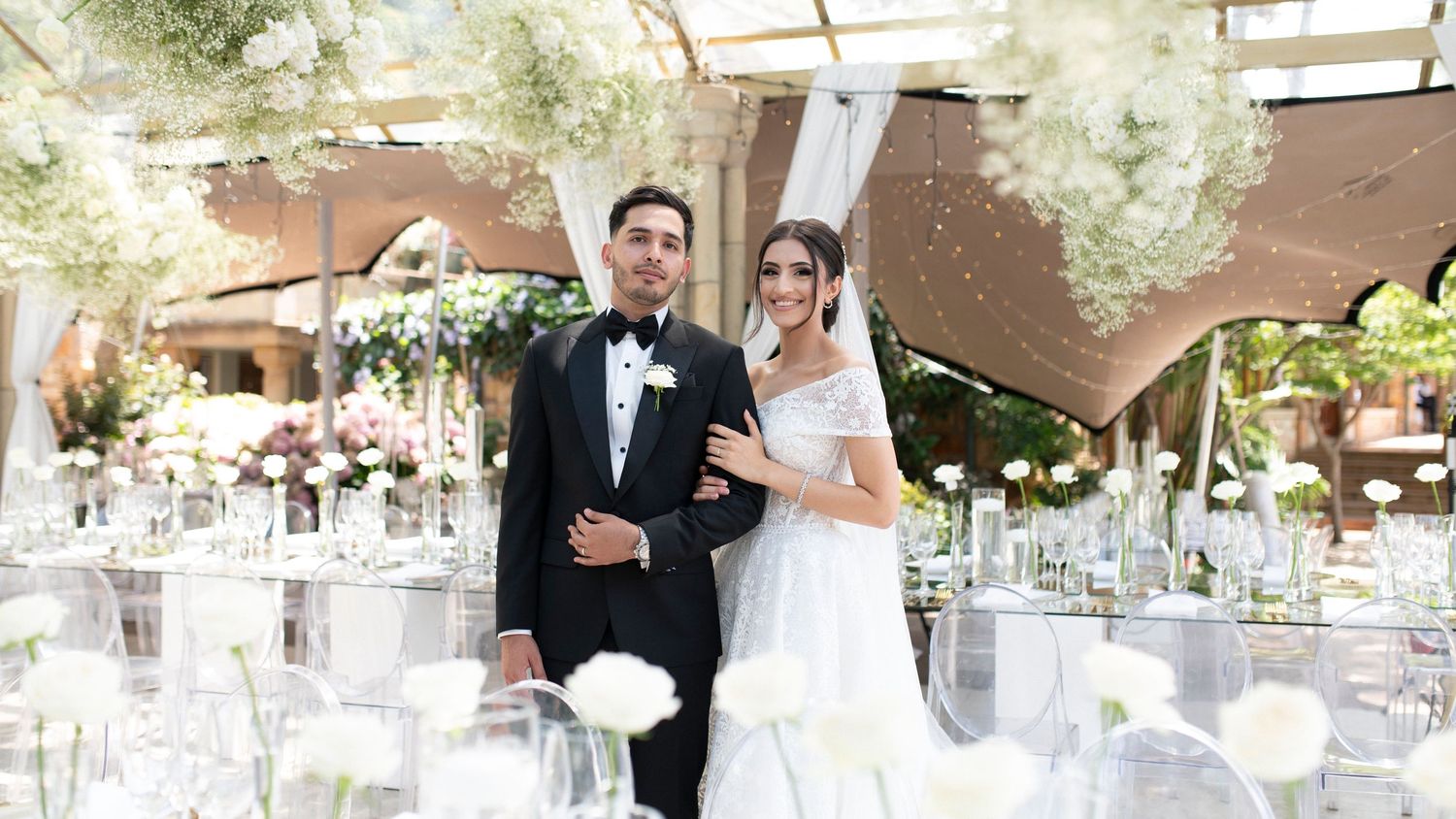 An outdoor wedding reception setup with white decor and floral arrangements under a tent structure.
