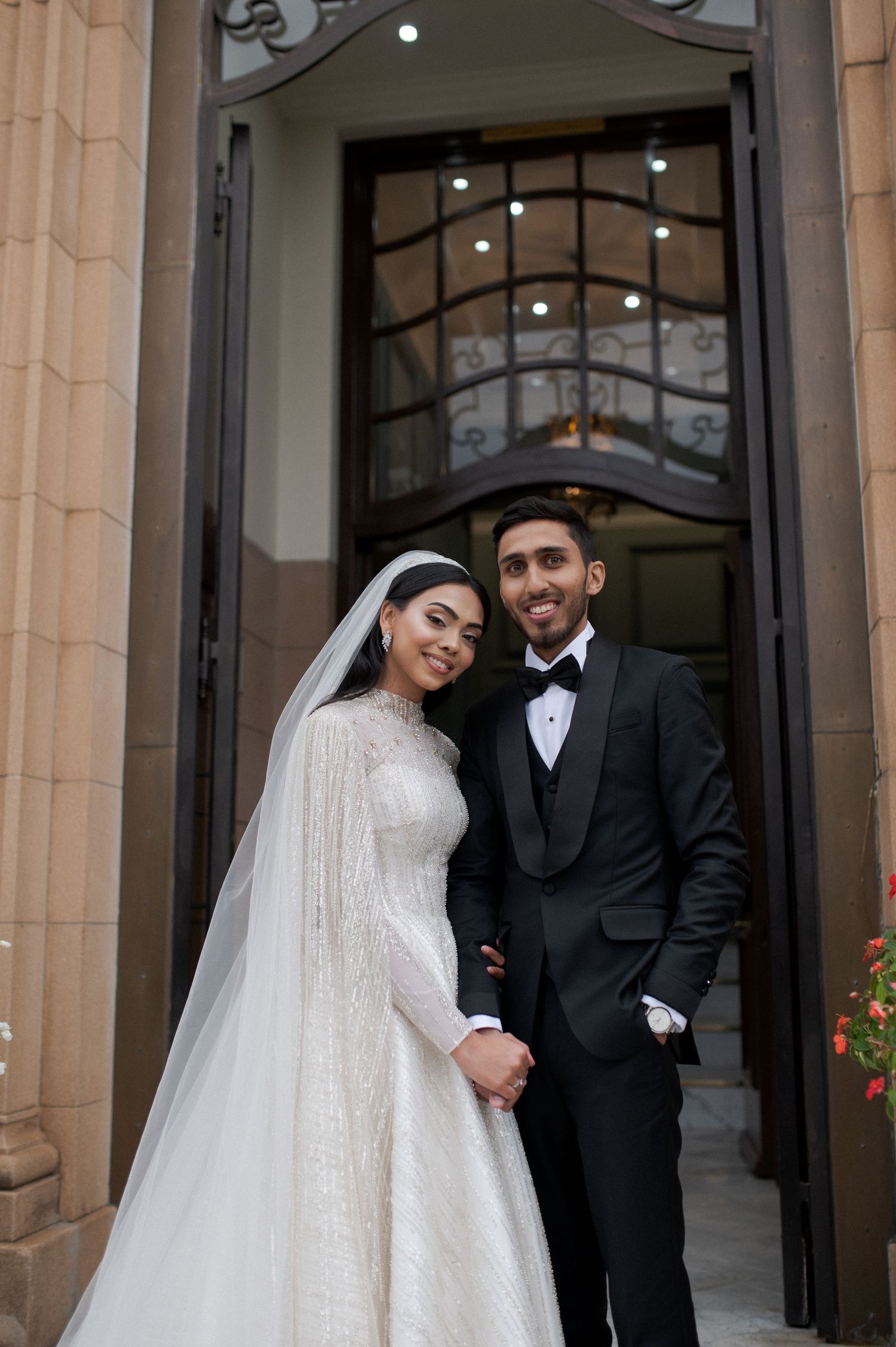 A wedding couple poses in front of an ornate doorway, the bride in white and groom in classic tuxedo.