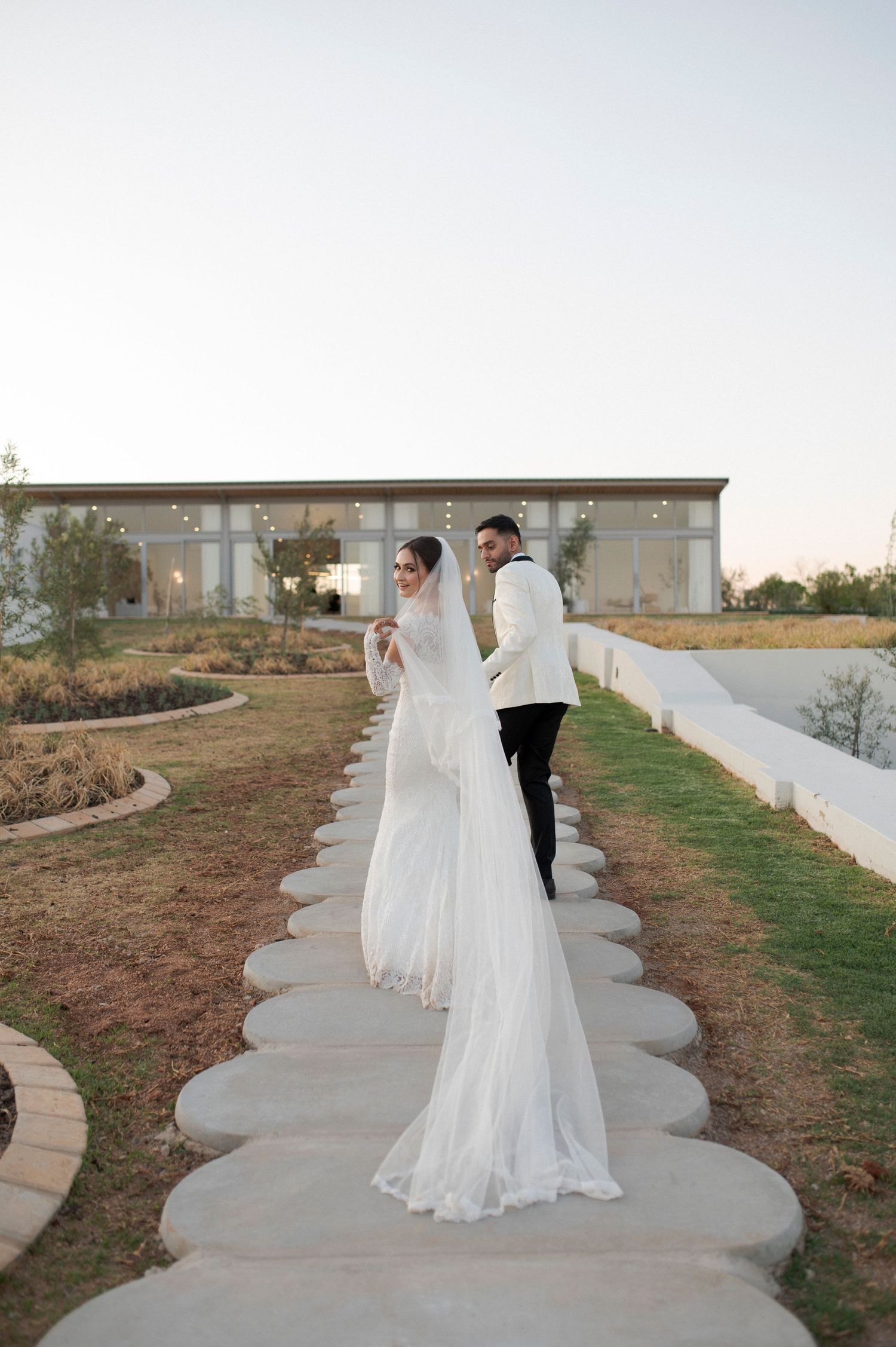 A wedding couple walks down a curved concrete path surrounded by landscaped grounds at sunset.
