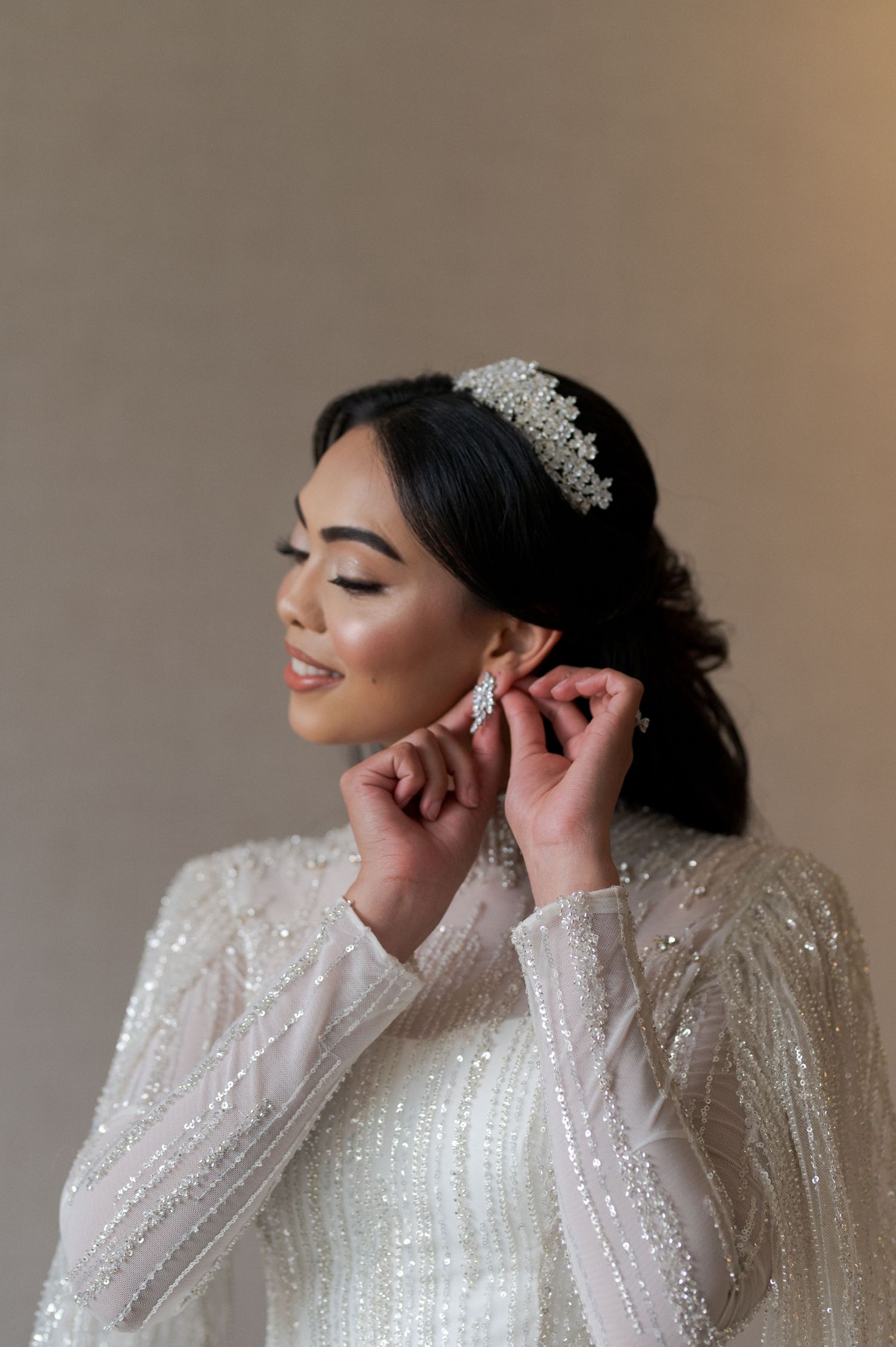 Bride wearing sparkly long-sleeve gown adjusts earrings while wearing crystal tiara.