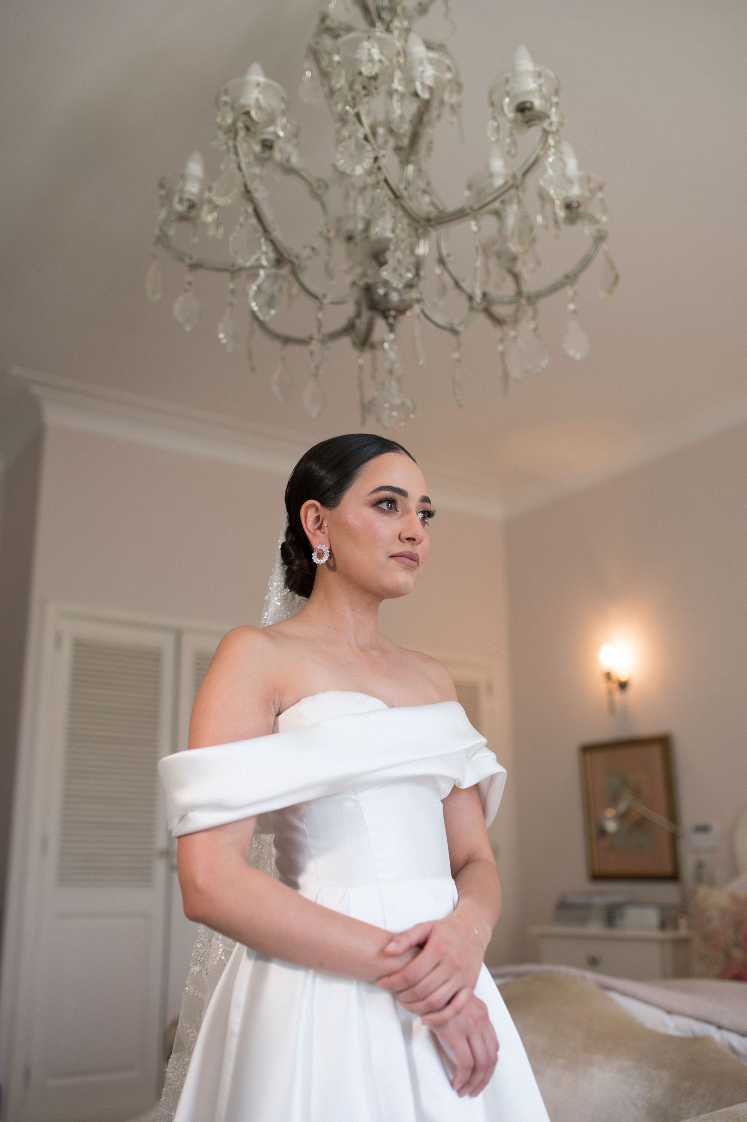 An elegant off-shoulder white wedding gown under a crystal chandelier.