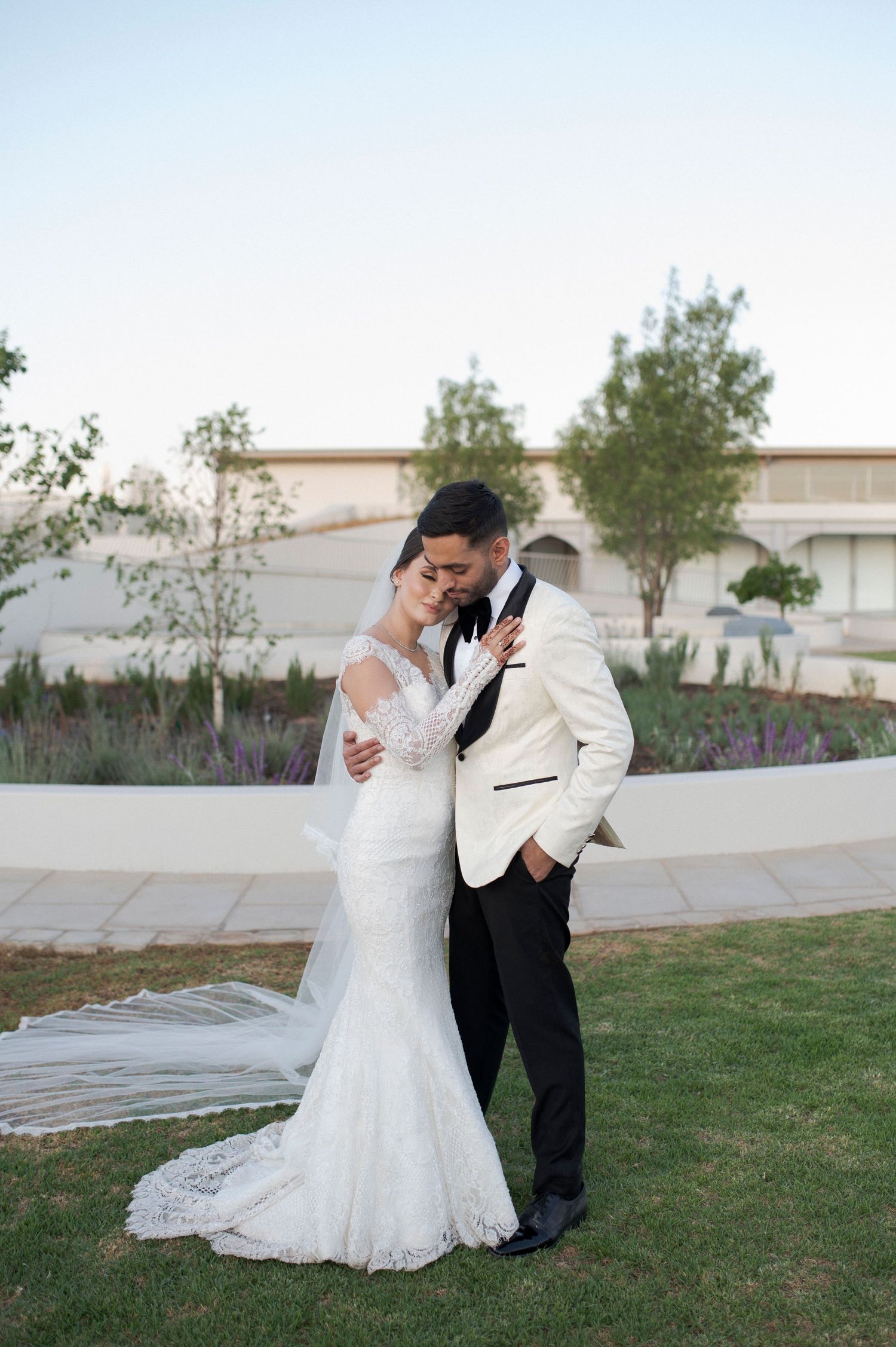 Romantic wedding couple embrace on manicured lawn with curved white wall backdrop.
