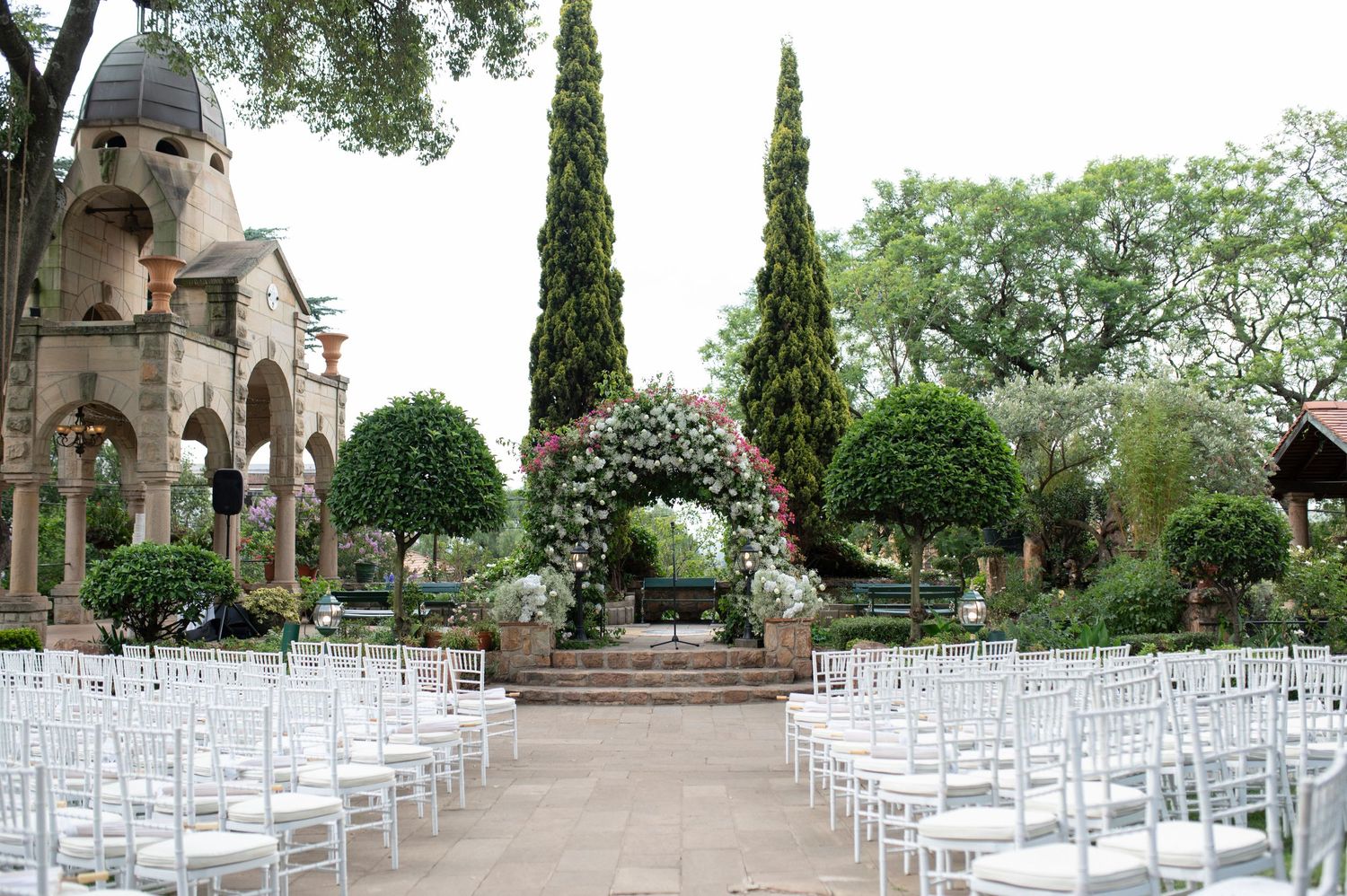 Outdoor wedding ceremony setup with white chairs and cypress trees leading to a stone archway.