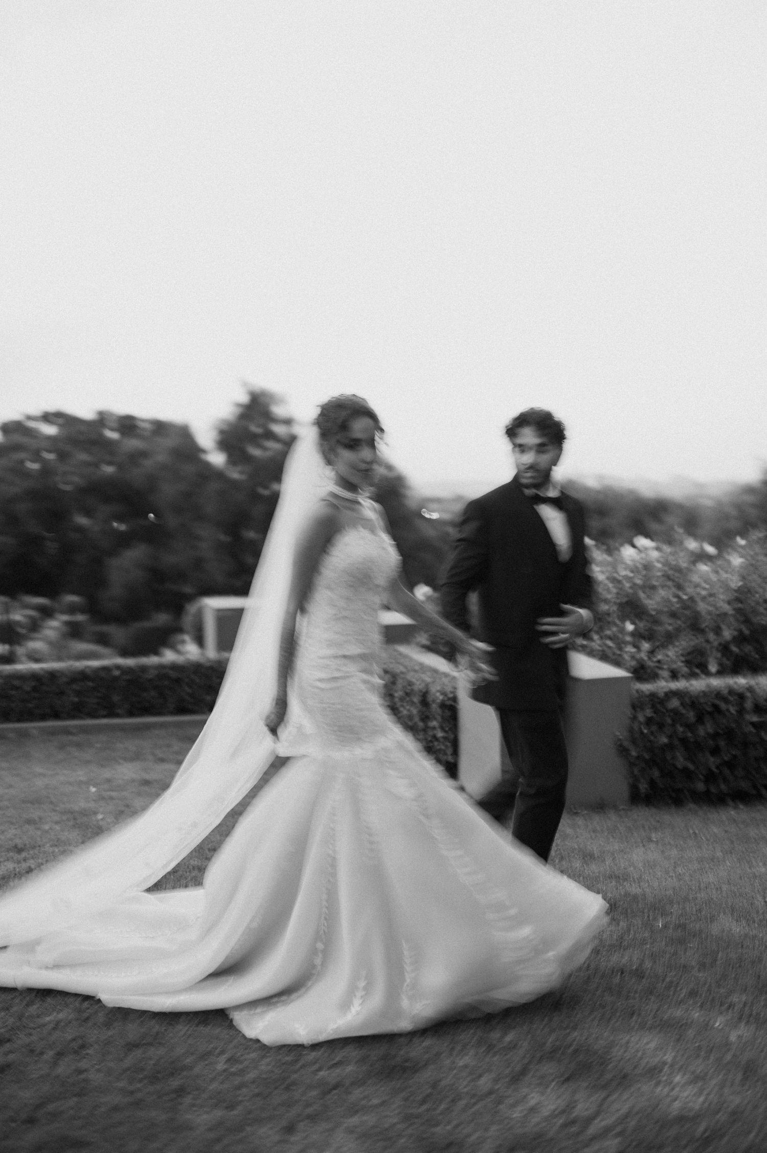 Black and white photo of newlyweds dancing in a garden with bride's dress flowing.